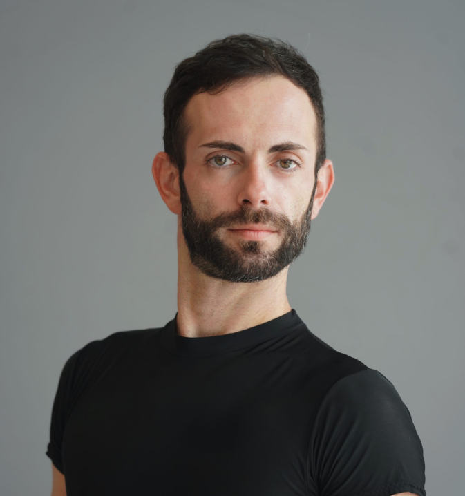 Picture of Patrick, man with dark hair and a beard, wearing a black shirt, looking at the camera against a gray background.
