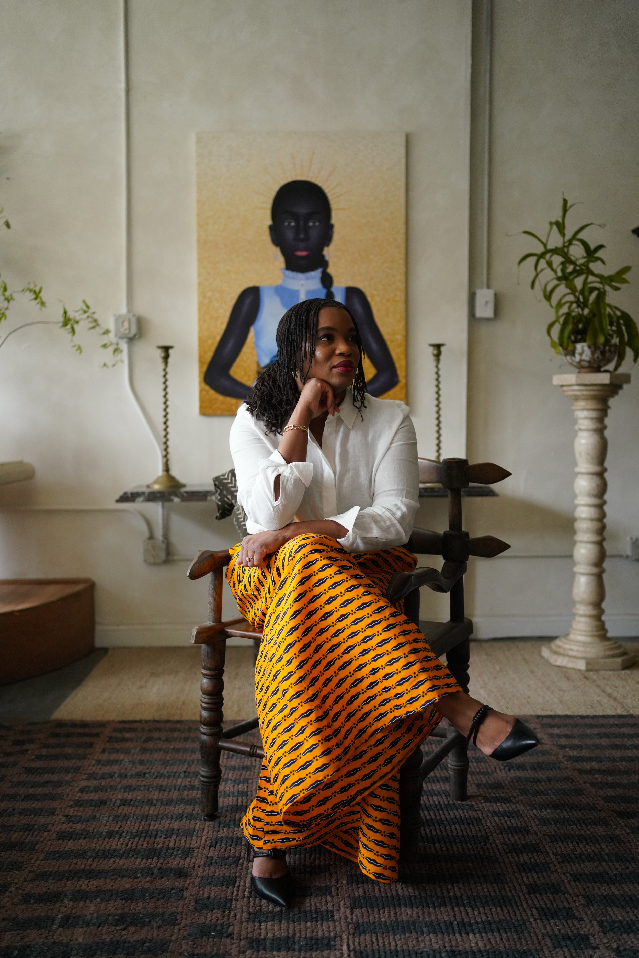 Ogechi in a white blouse and colorful patterned Ankara pants sits on an African wooden chair in a room with artistic decor and a painting on the wall behind her. The room features plant decorations and sophisticated lighting.