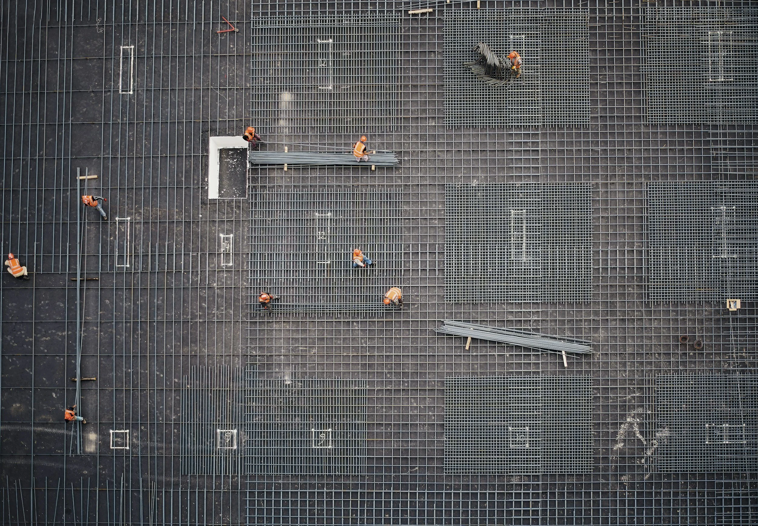 Top-down view of construction workers installing metal grates on a dark cement floor, with some workers in orange safety vests and helmets.