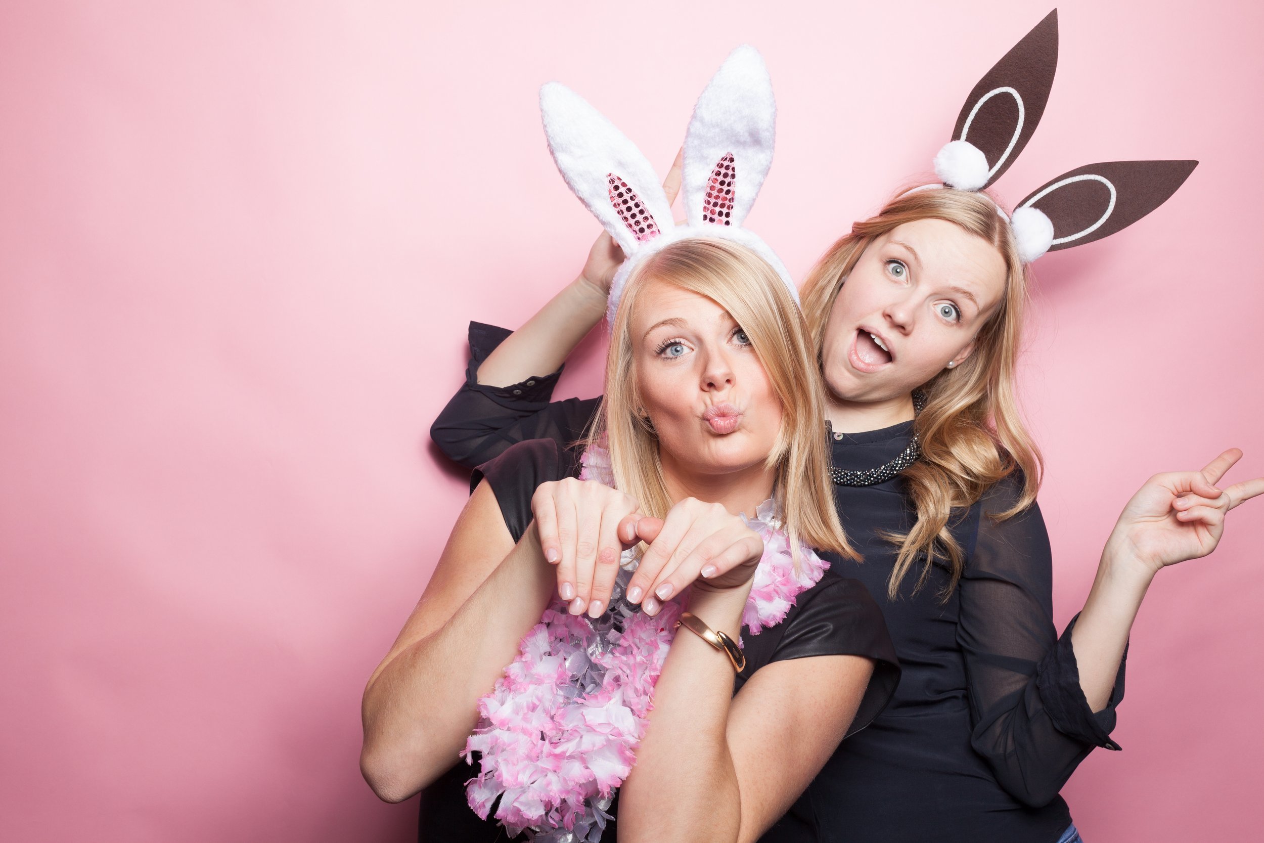 Two women posing playfully in front of a pink background, wearing bunny ears and making fun expressions.