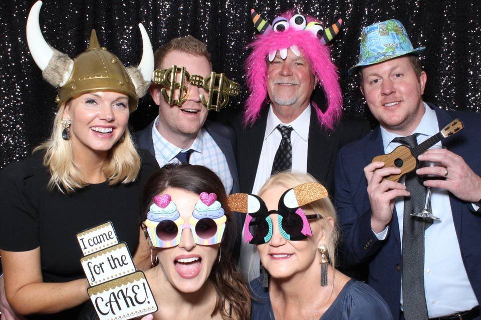 Group of people at a party wearing funny costumes and accessories, including Viking helmet, novelty glasses, and colorful hats, posing in front of a glittery backdrop, with a sign saying "I came for the cake!"