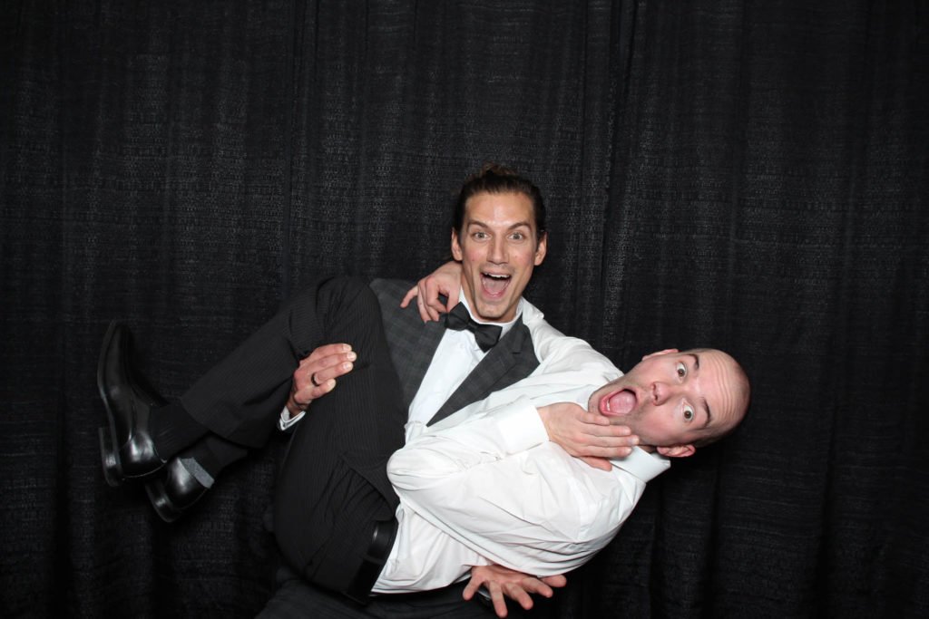 Two men in suits posing playfully; one is being carried by the other against a black curtain backdrop, both with surprised expressions.