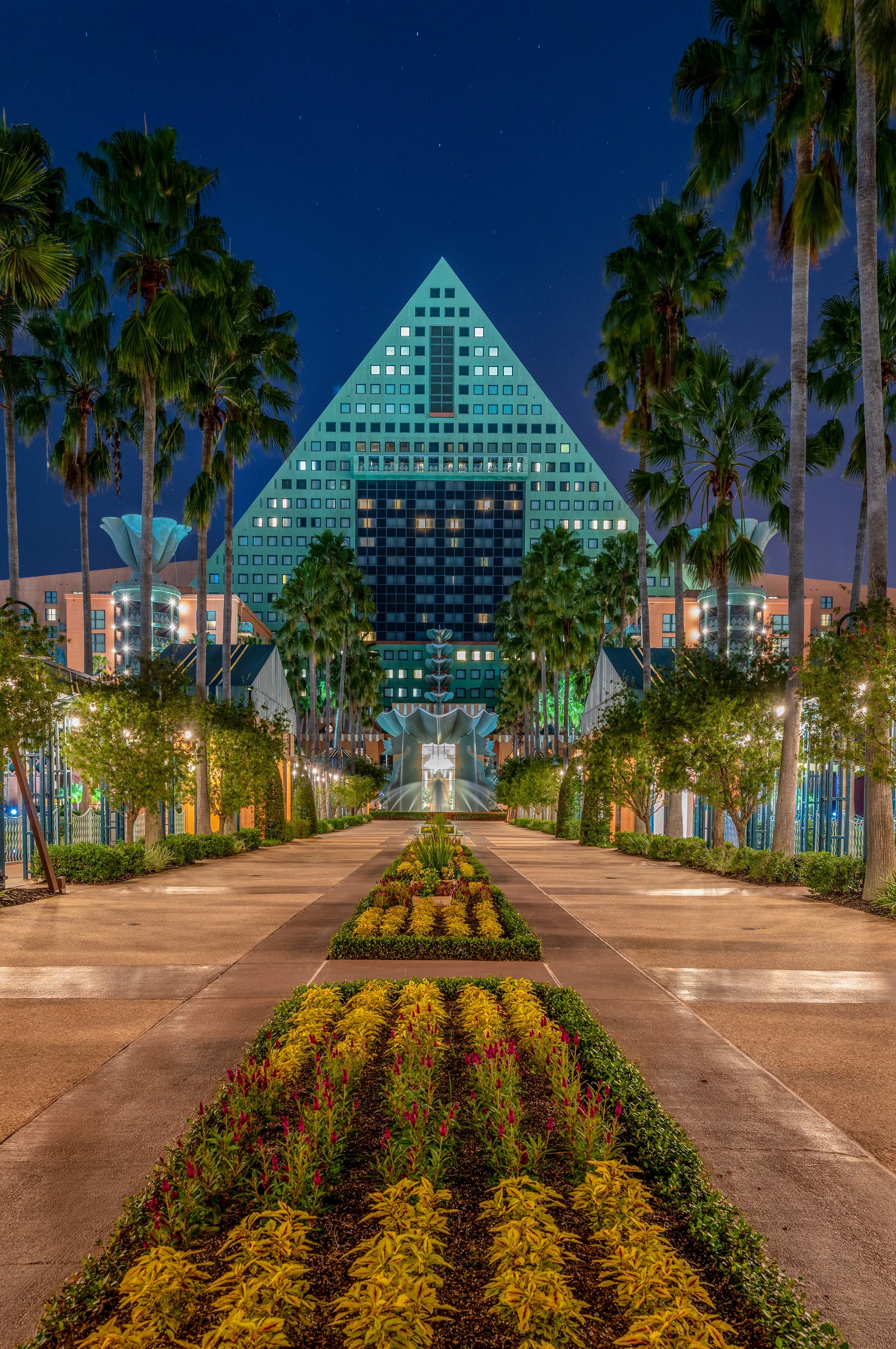 Night scene of a brightly lit, modern pyramid-shaped hotel with palm trees lining a walkway leading to it, and landscaped flower beds in foreground.