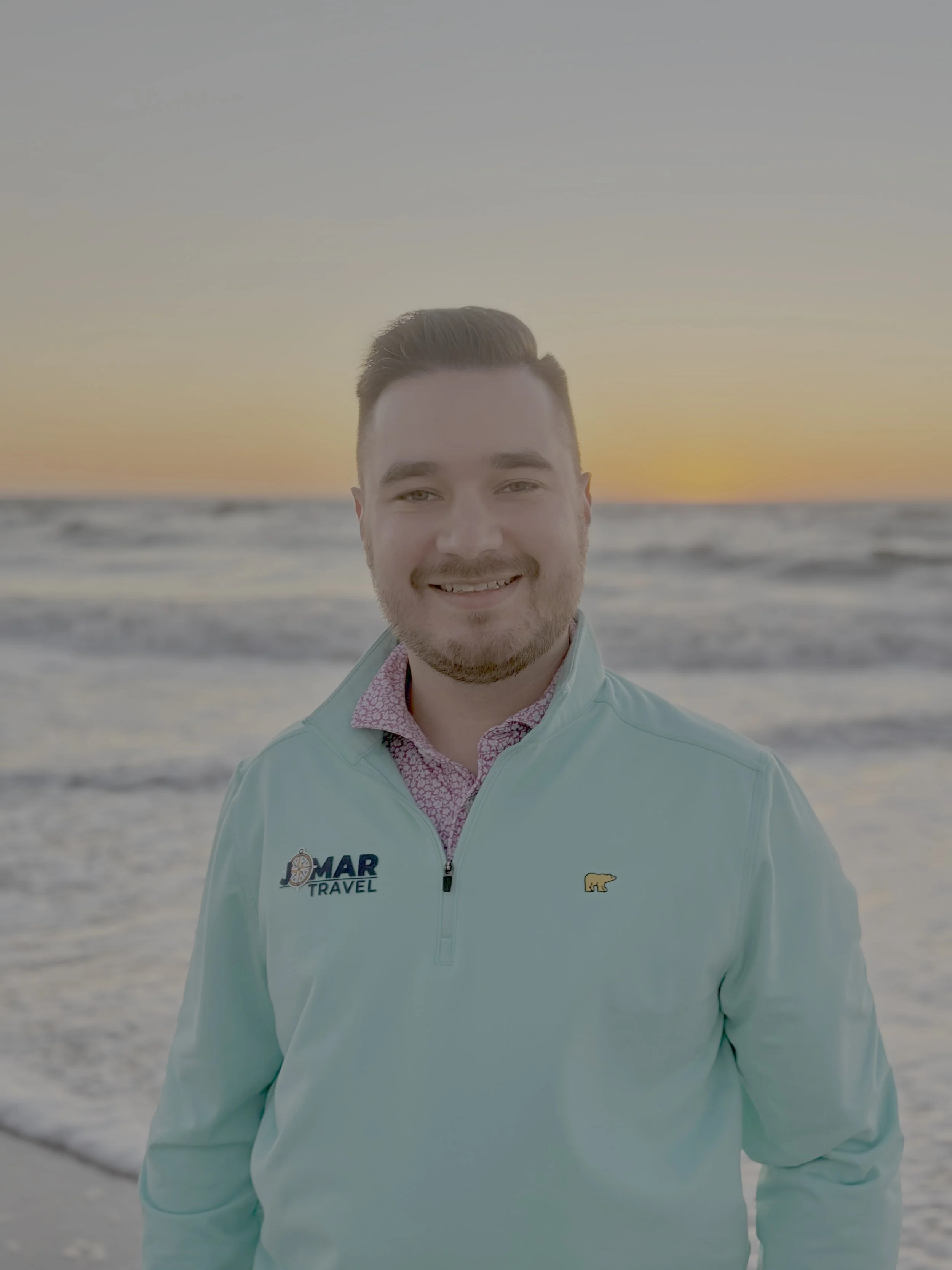A man smiling at the beach during sunset, wearing a light blue jacket with a travel logo and a yellow bear logo.