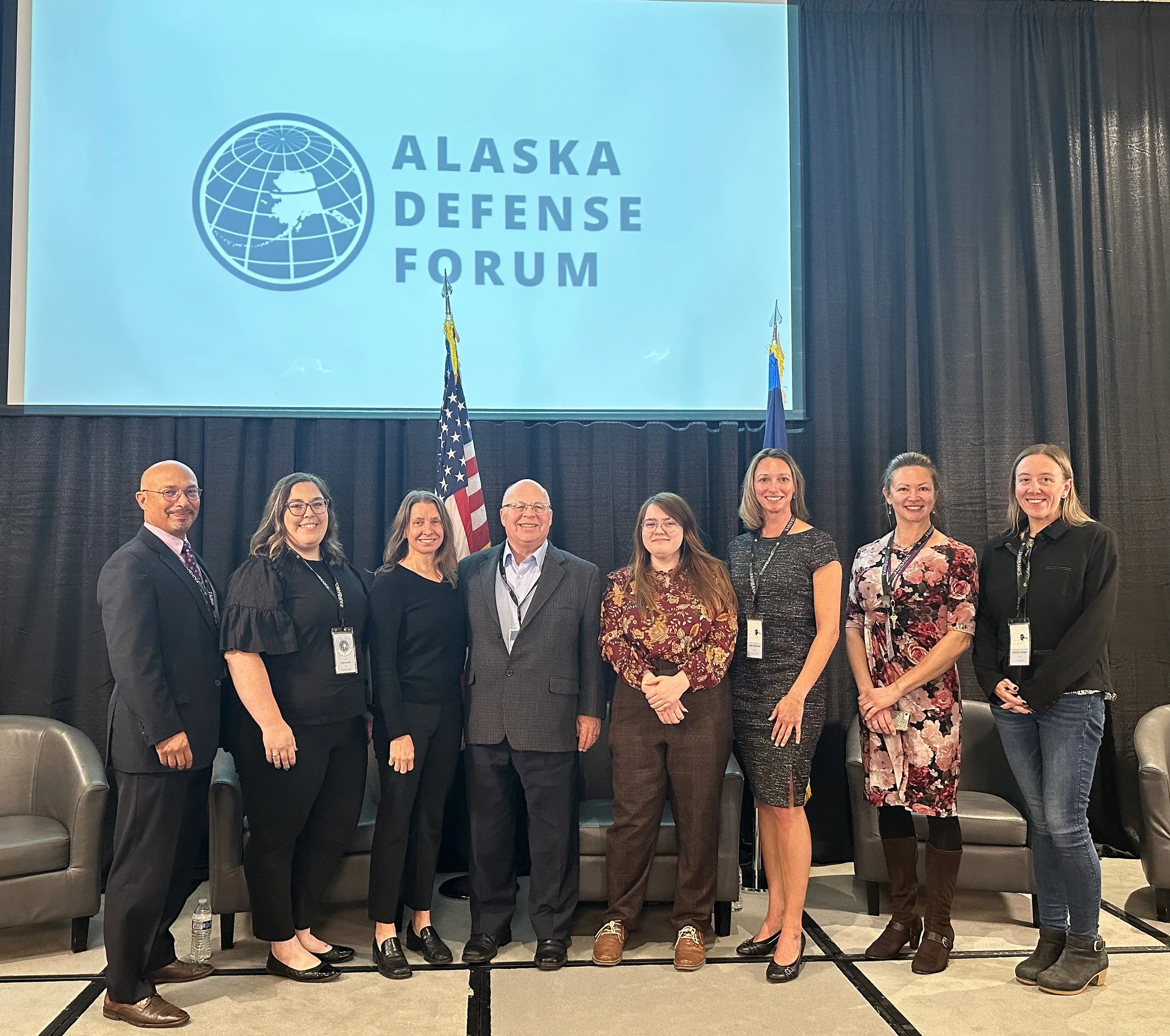 Jennifer Thompson and a group of members posing in front of an Alaska Defense Forum sign.
