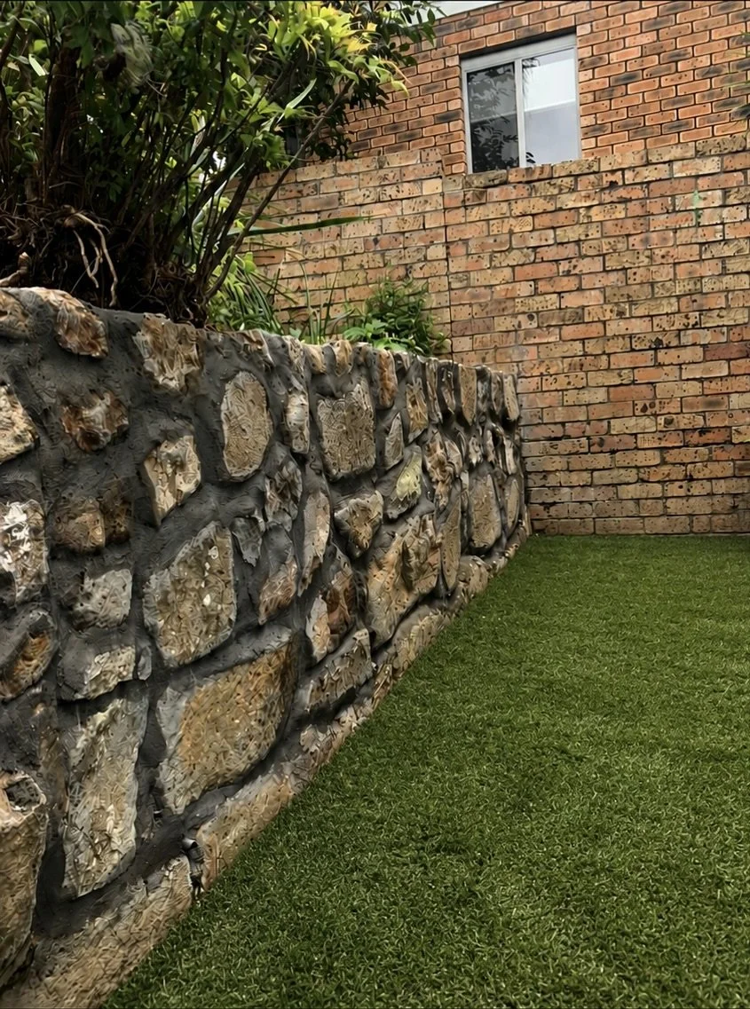 A stone retaining wall with embedded shells and a lush green lawn in front. There are plants and a brick house with a window in the background.
