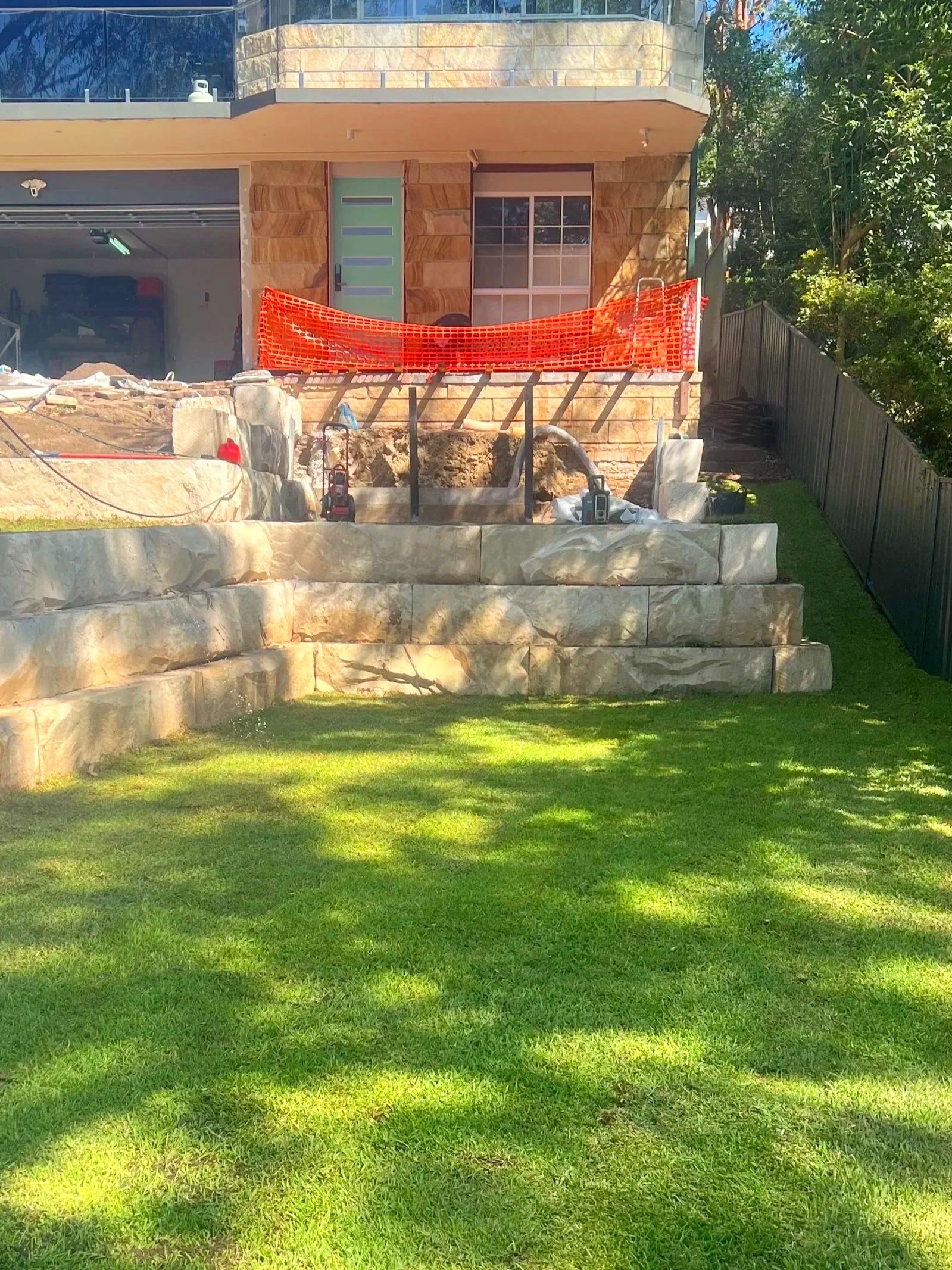 Construction site in backyard with stone retaining wall, green grassy yard, and house in the background.