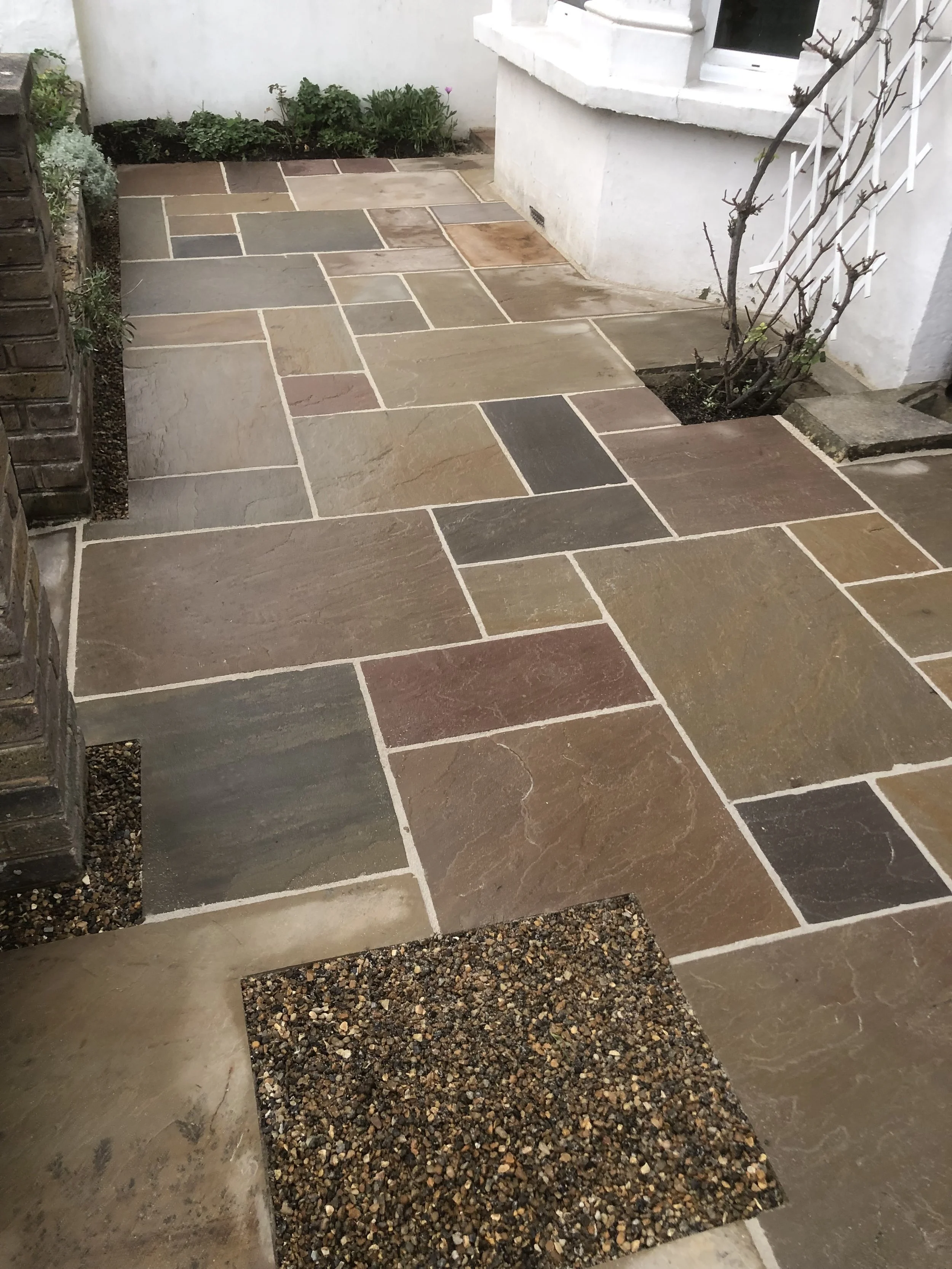 Colorful stone patio with a small square gravel section, adjacent to a white house with a window and plants.