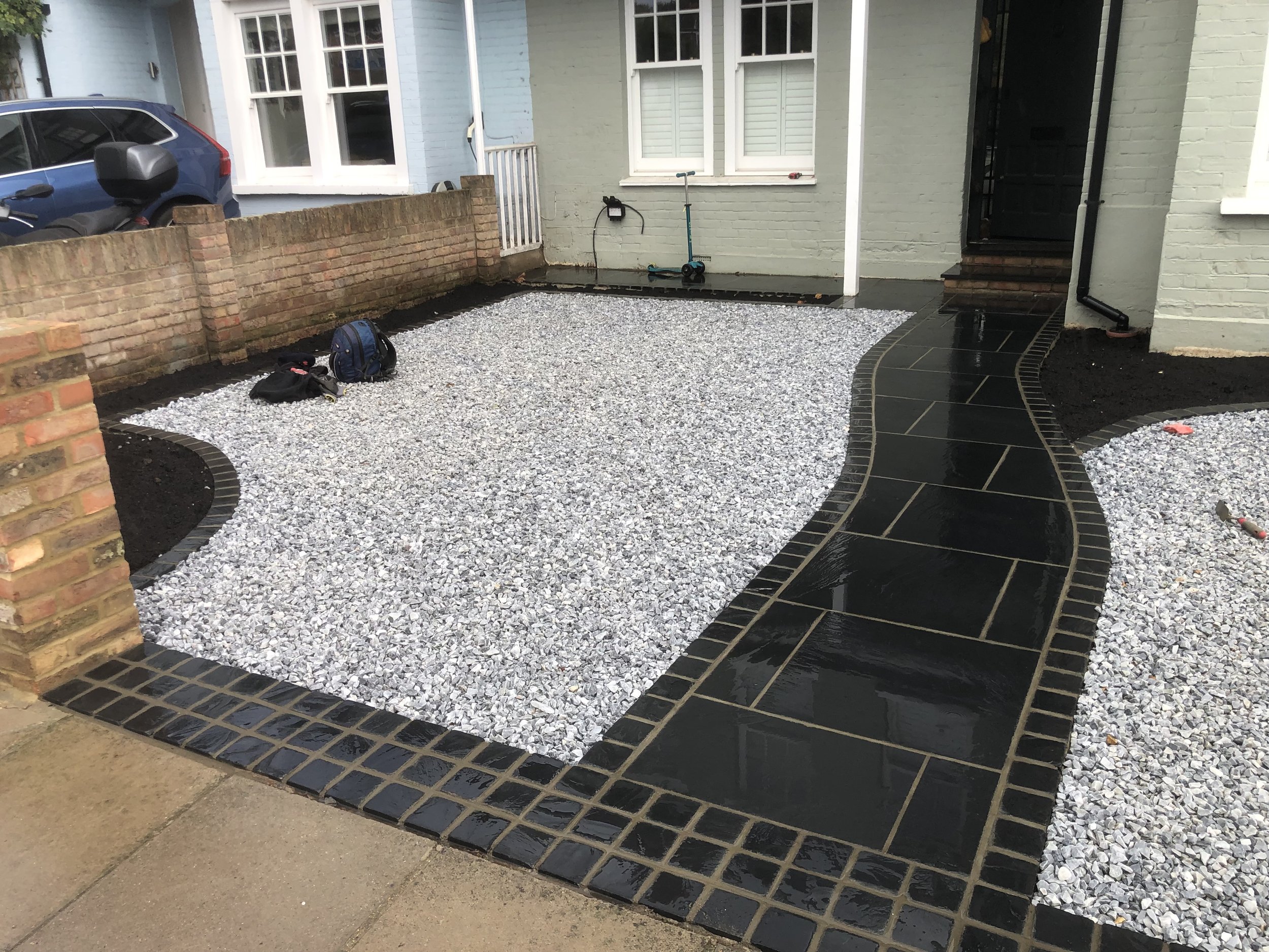 Front yard with black paved pathway, white gravel, and brick edging. A house with light green painted brick exterior and white window shutters in the background.