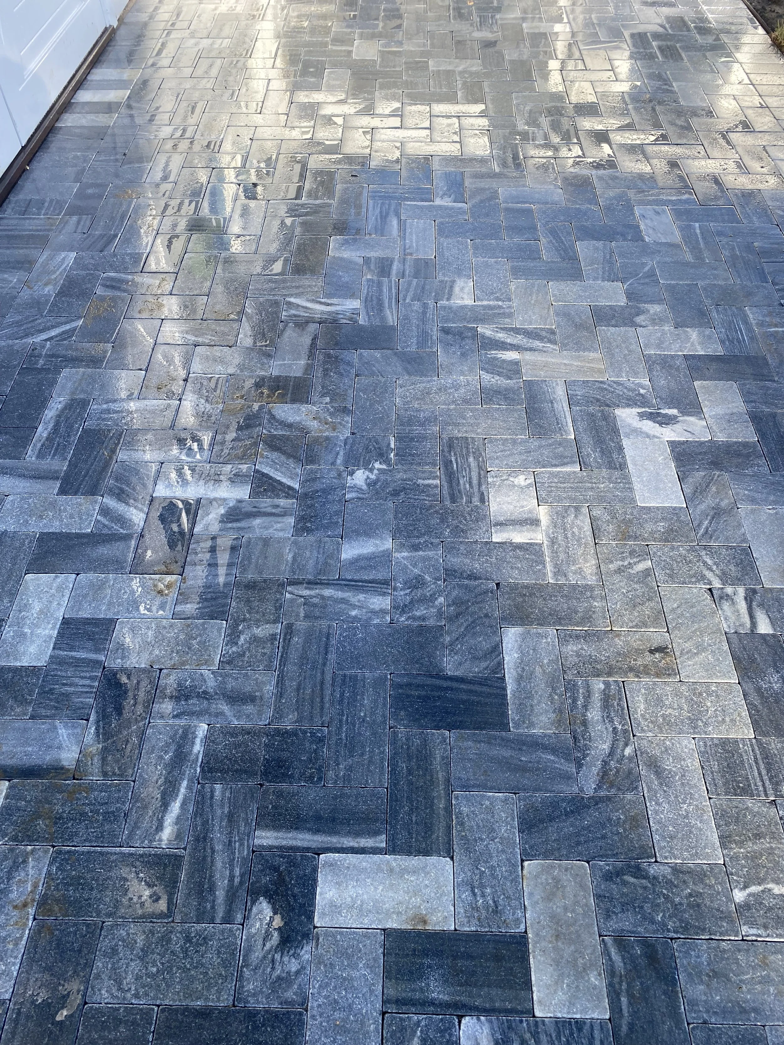 A paved outdoor area with grey stone pavers laid in a herringbone pattern, with some wet spots and reflections visible.