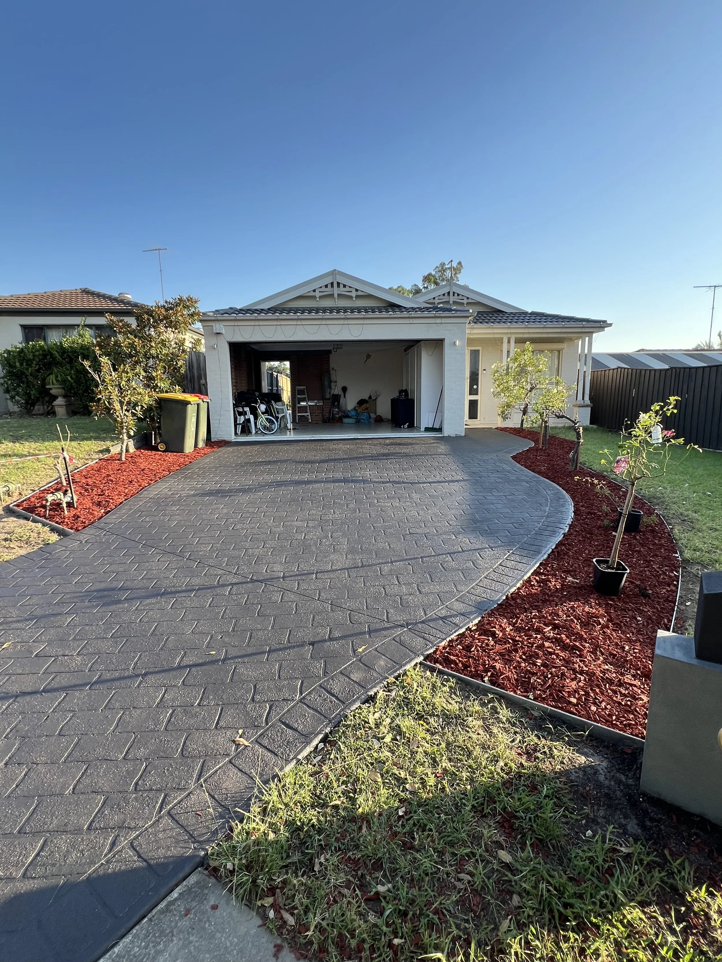 A house with a brick open garage, a curved paved driveway, small trees and landscaped garden beds with red mulch.