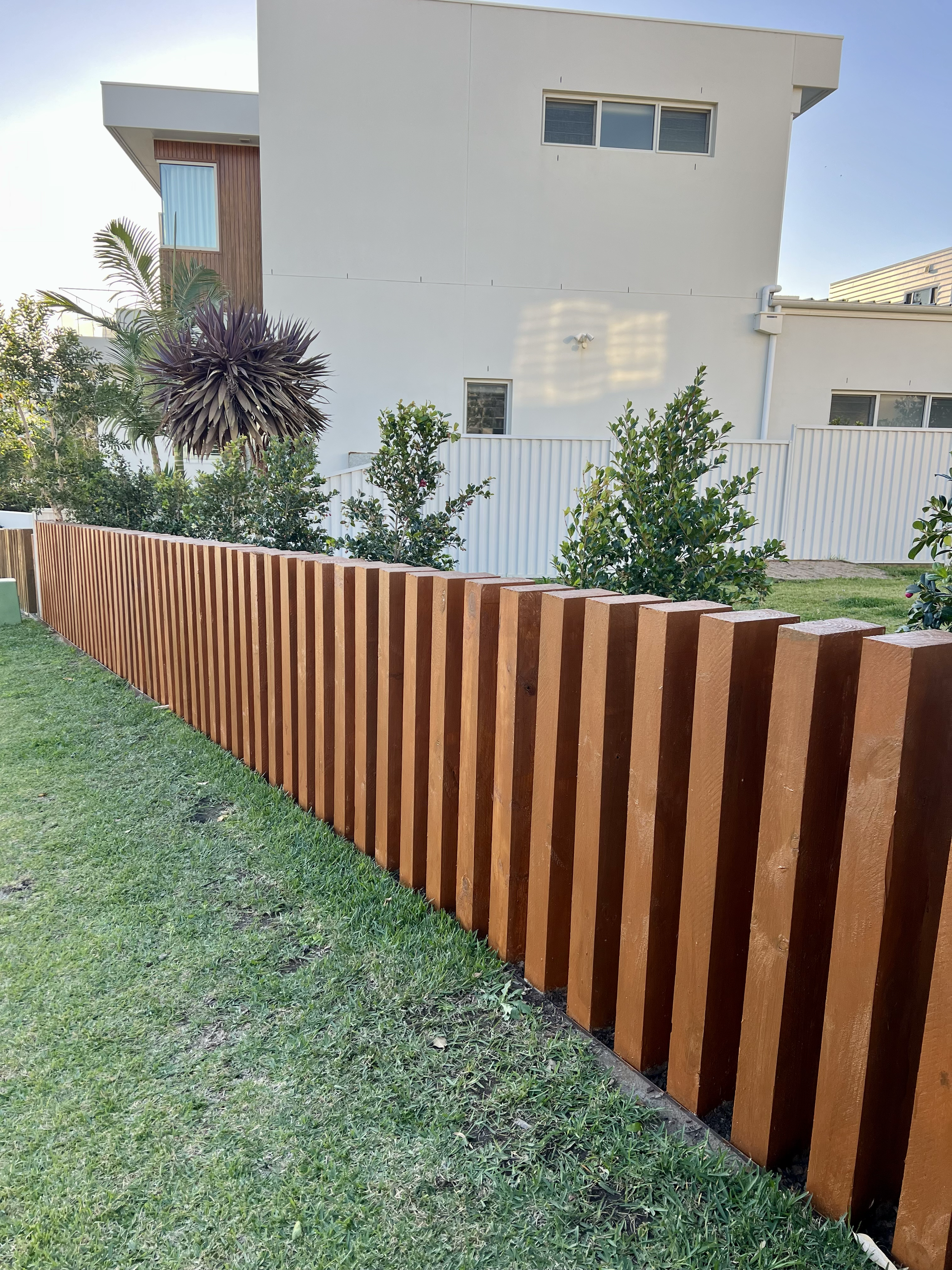 Residential backyard with a red wooden fence, lush green grass, and various shrubs and trees, with a modern white house featuring small windows and a small balcony in the background.
