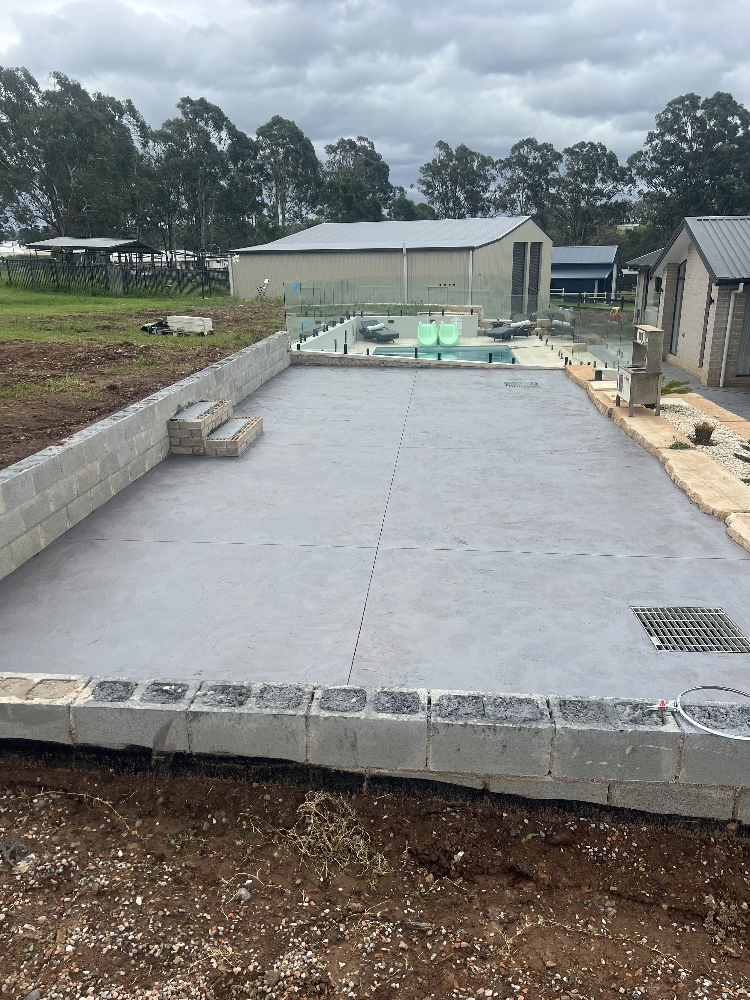 Concrete patio in a backyard with a partially built brick wall and steps, surrounded by dirt, with a small pool and patio furniture in the background under a cloudy sky.