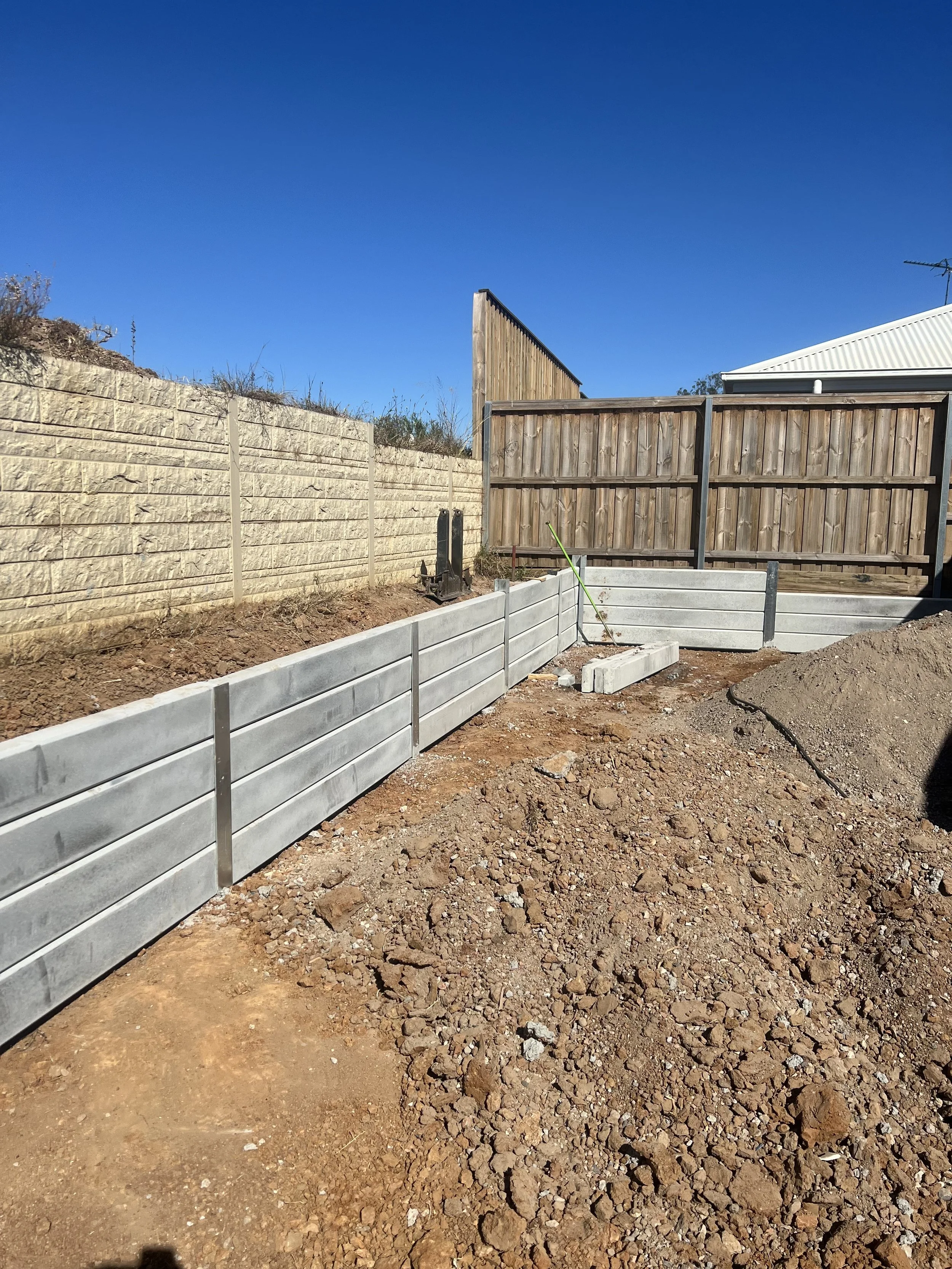 Construction site with a newly built concrete retaining wall, dirt ground, wooden fence, and a clear blue sky.