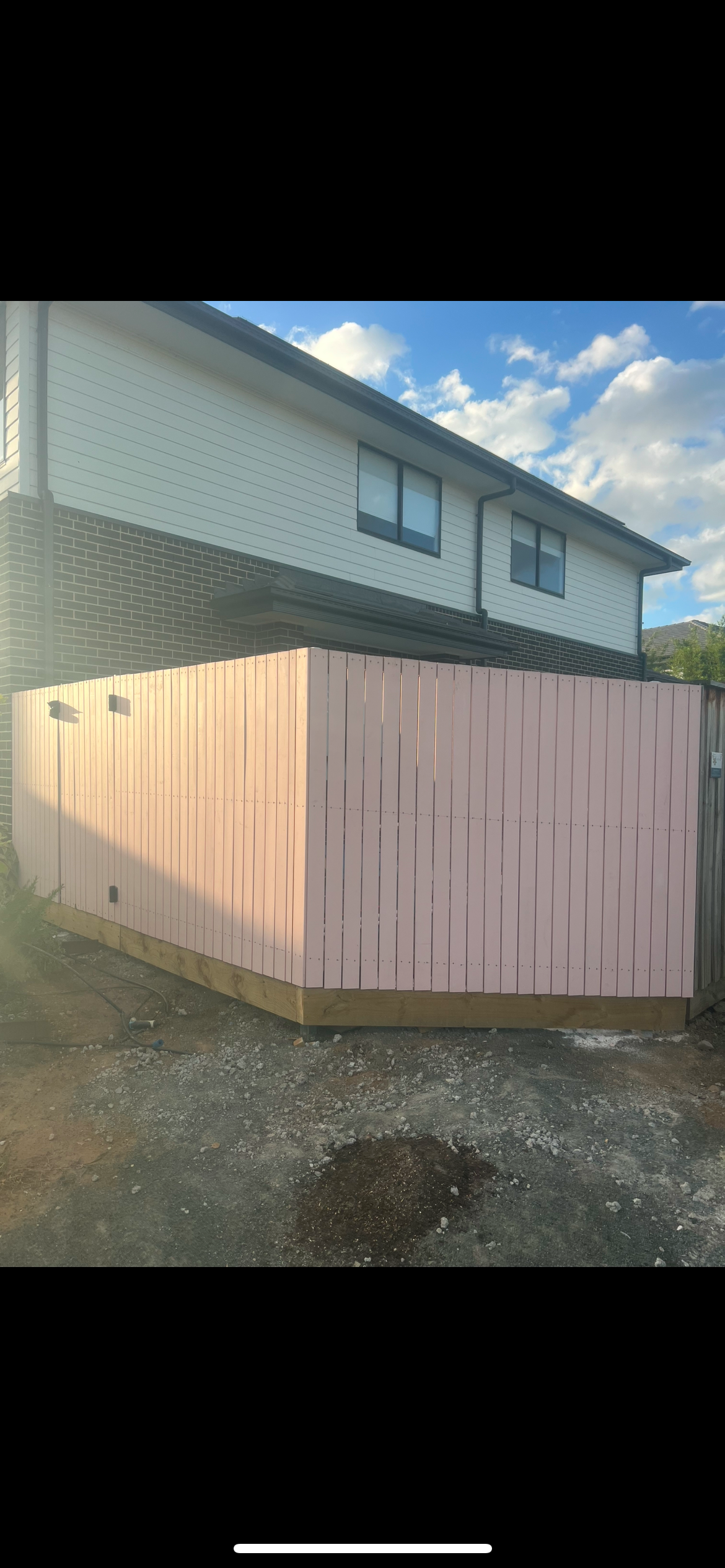A two-story house with black window frames and white siding, featuring a brown brick lower level. In front, there is a pink wooden fence with vertical slats and a wooden bottom trim, surrounding a small yard with dirt and gravel. The sky is partly cl