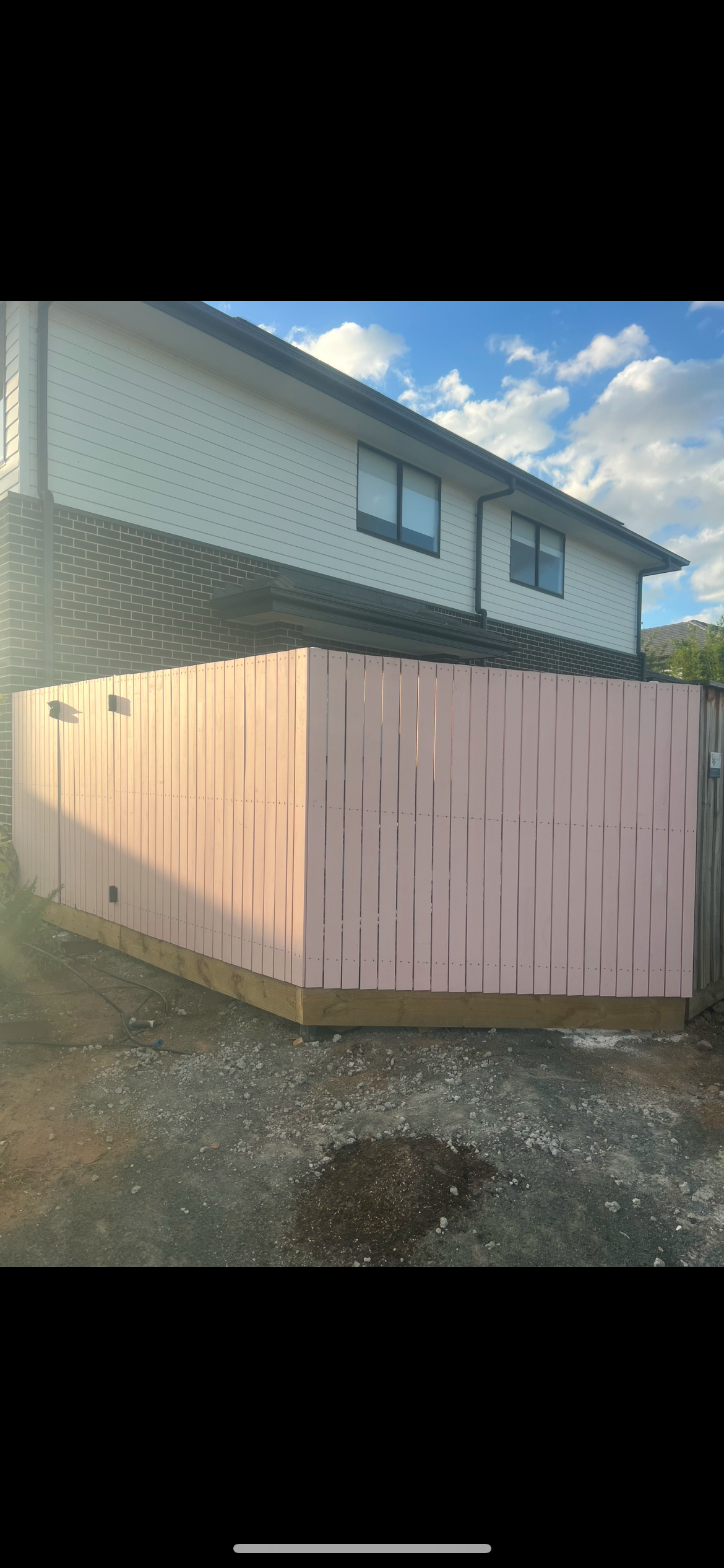 A light pink wooden privacy fence around a backyard area, with part of a two-story house visible in the background, under a partly cloudy sky.