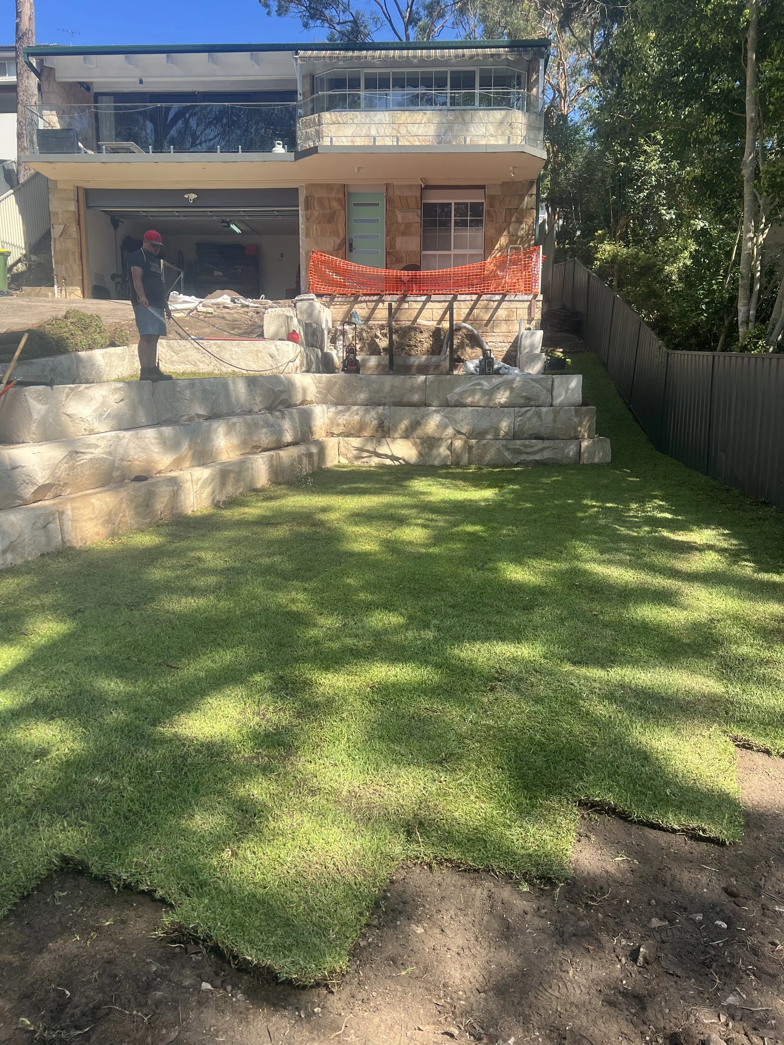 Backyard with a green lawn, stone patio, and a house under construction, with workers and construction materials present.