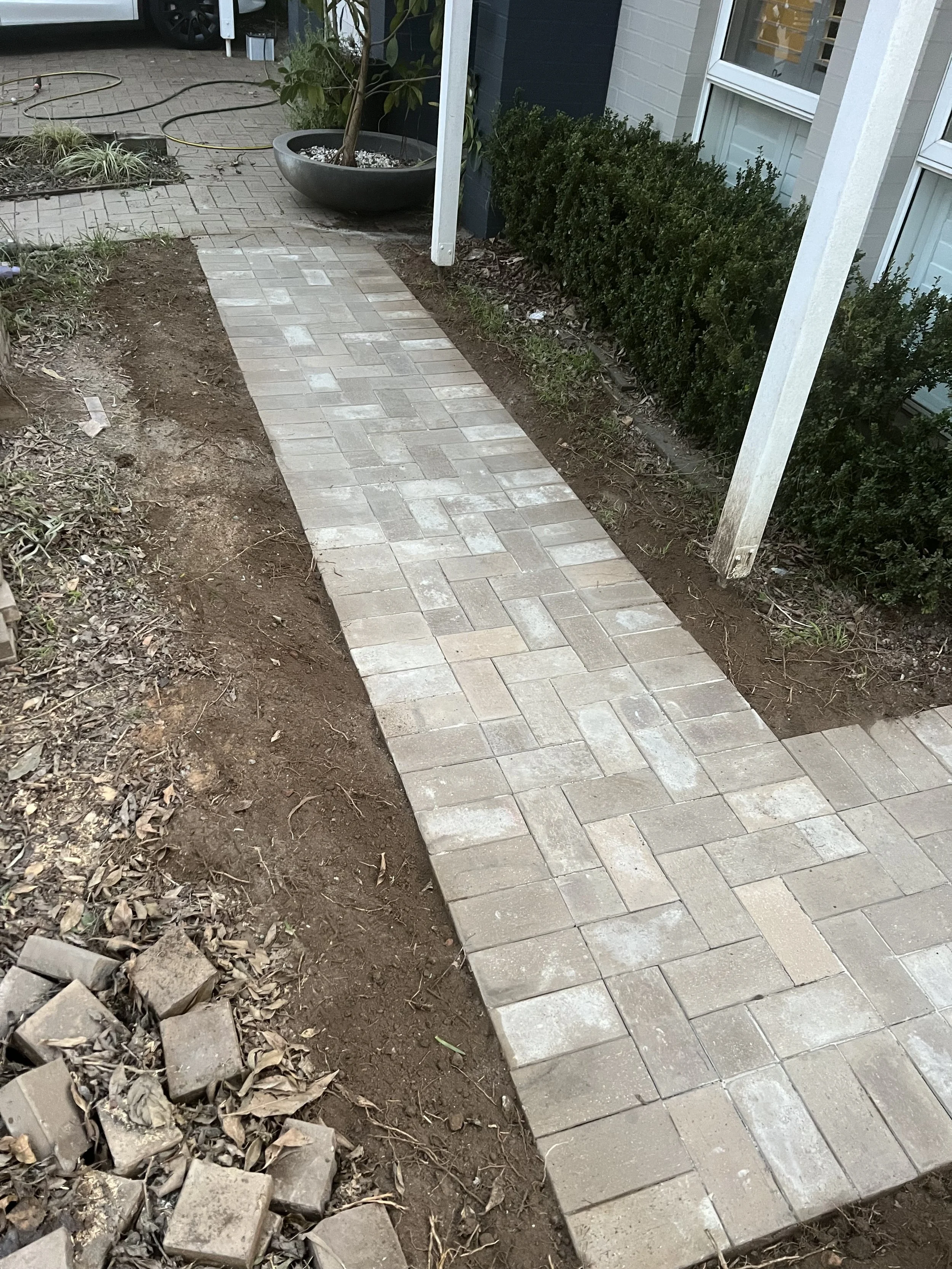 Newly installed brick walkway next to a house with bushes, a potted plant, and some dirt and debris on the side.