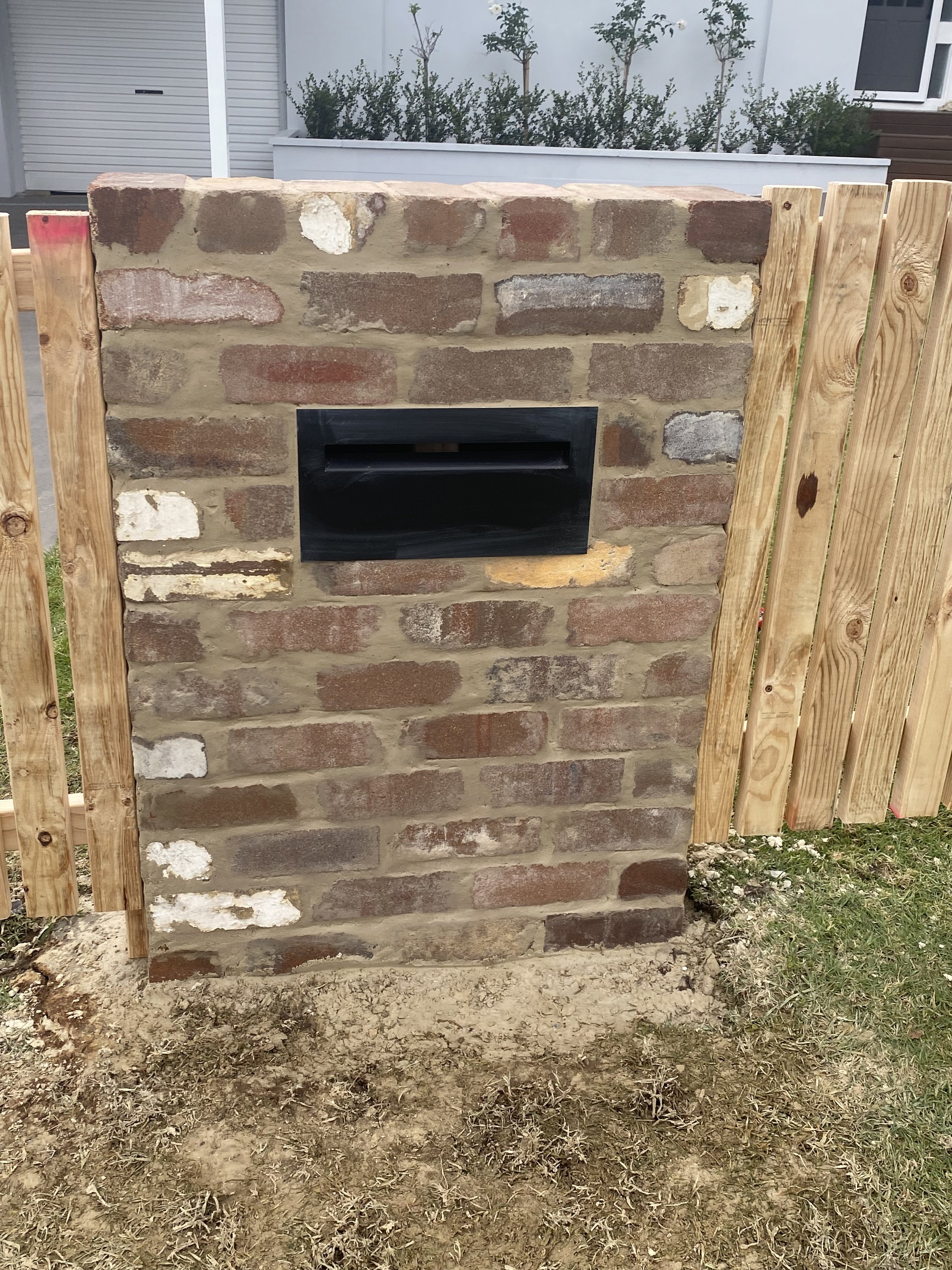 A brick wall with a black mailbox in the center, flanked by wooden posts and fence panels. Behind the wall, there is a white planter with shrubs and a house with a garage door.