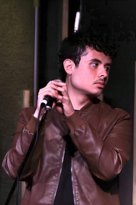 A young man with curly dark hair wearing a brown leather jacket holding a microphone with his right hand, looking to his right in a dimly lit indoor setting.