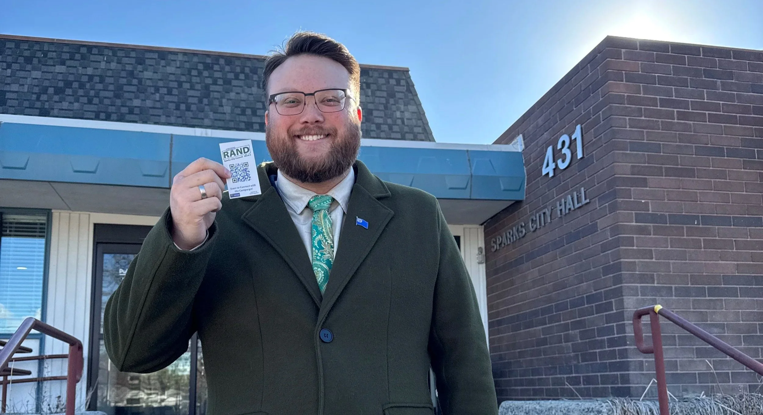 Hunter Rand stands in front of Sparks City Hall holding up a business card.