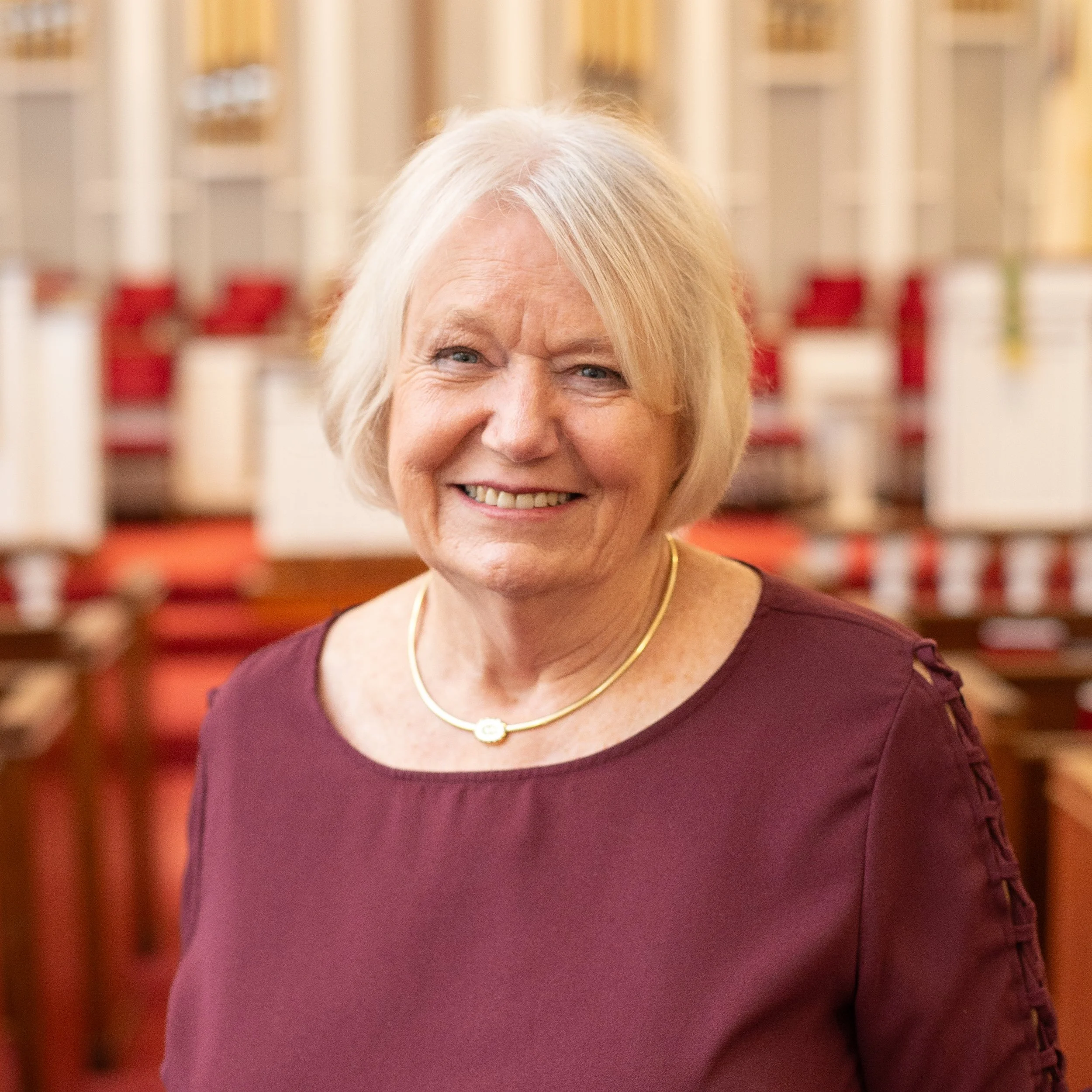 A woman wearing a light blue blouse standing inside a church with wooden pews and stained glass windows in the background.