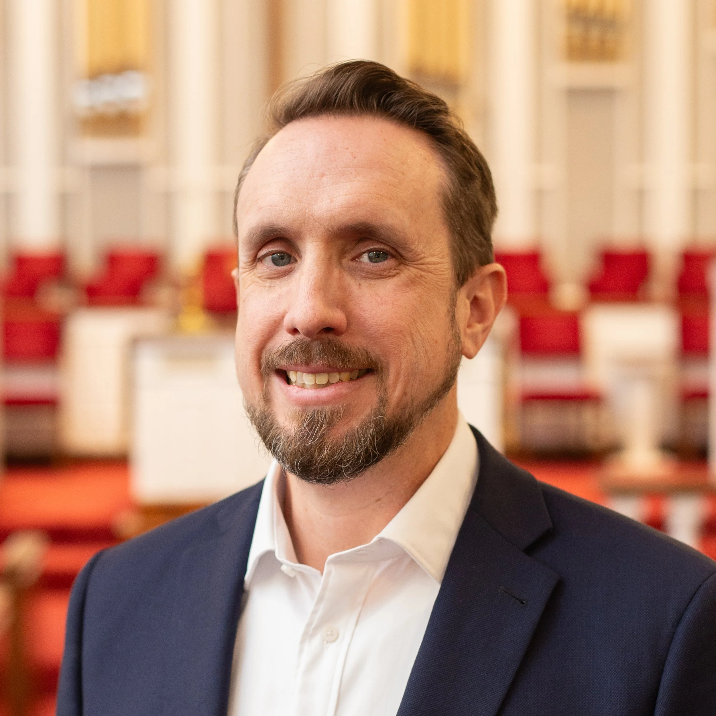 A man with a beard wearing a blue suit and gray shirt standing indoors with stained glass windows in the background.