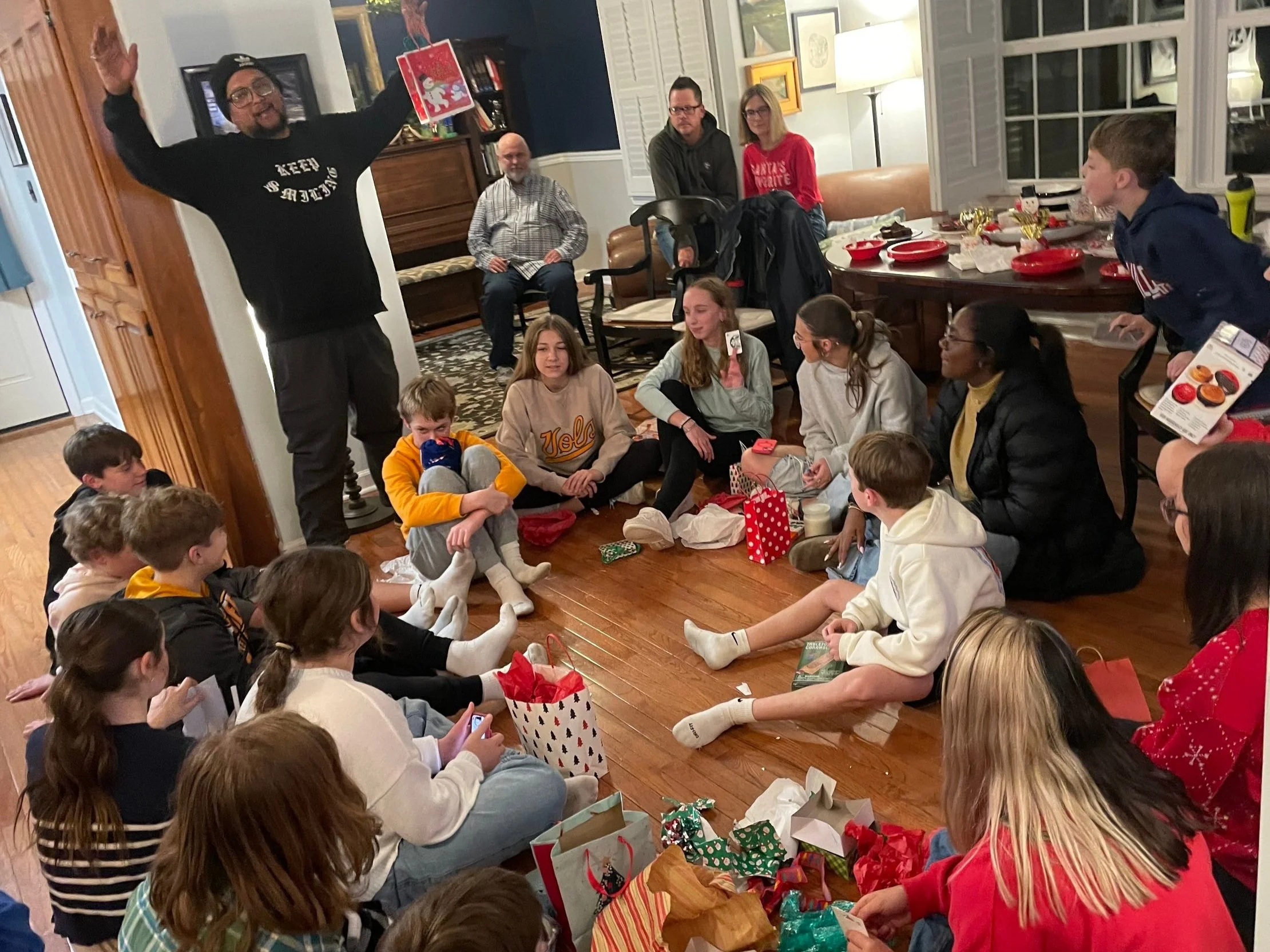 A group of people, including children and adults, sitting on a wooden floor and participating in a holiday gift exchange. There are gift bags and wrapping paper scattered around. One person is standing with arms raised, holding a gift bag. The room has a festive atmosphere with a table of red plates in the background.