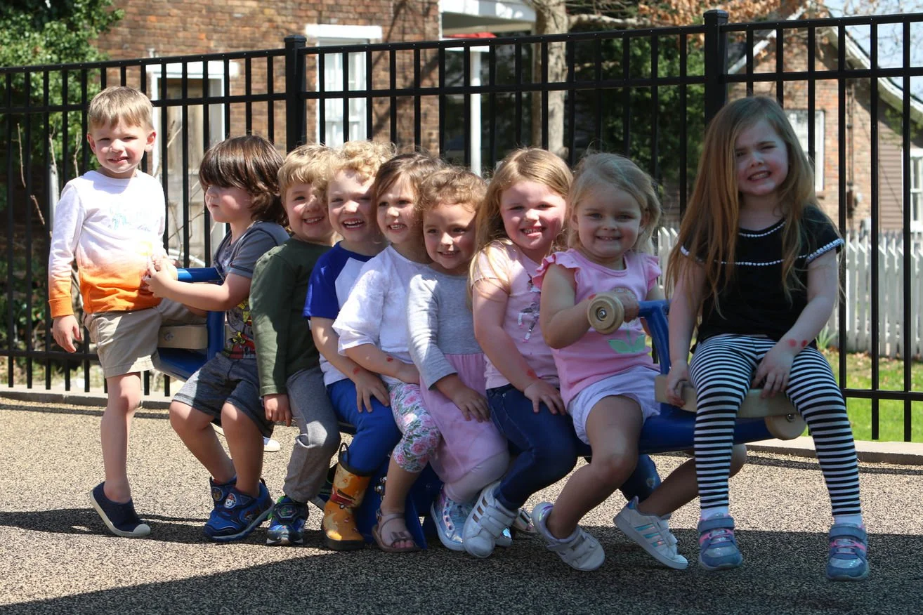 Group of children sitting on a playground seesaw, smiling in a sunny outdoor setting.