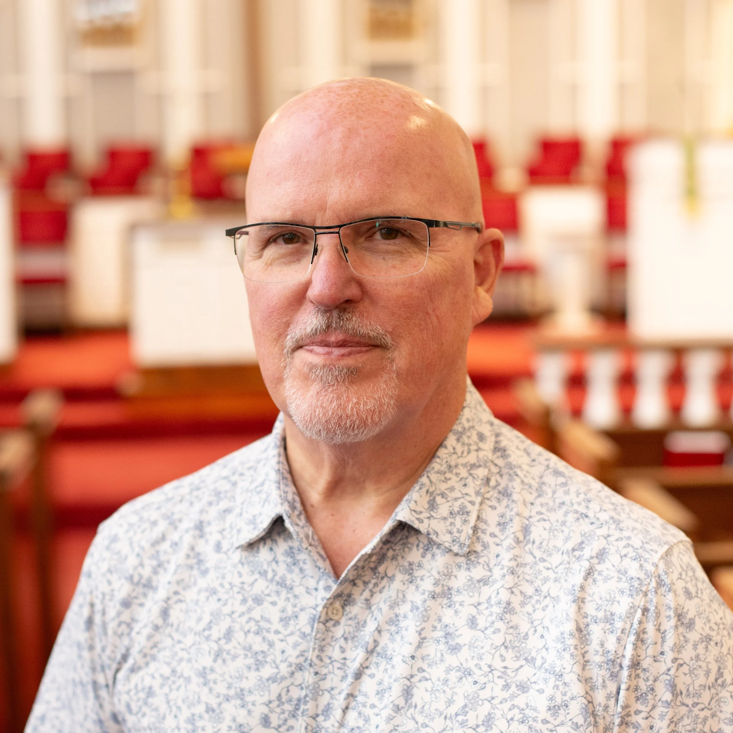 Bald man with glasses and a light patterned shirt smiling, standing in front of a bookshelf with decorative items.