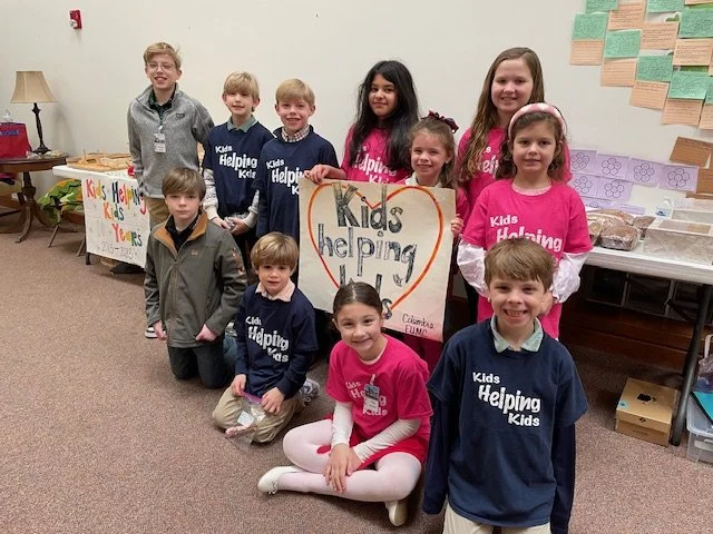 Group of children smiling and holding signs reading "Kids Helping Kids."