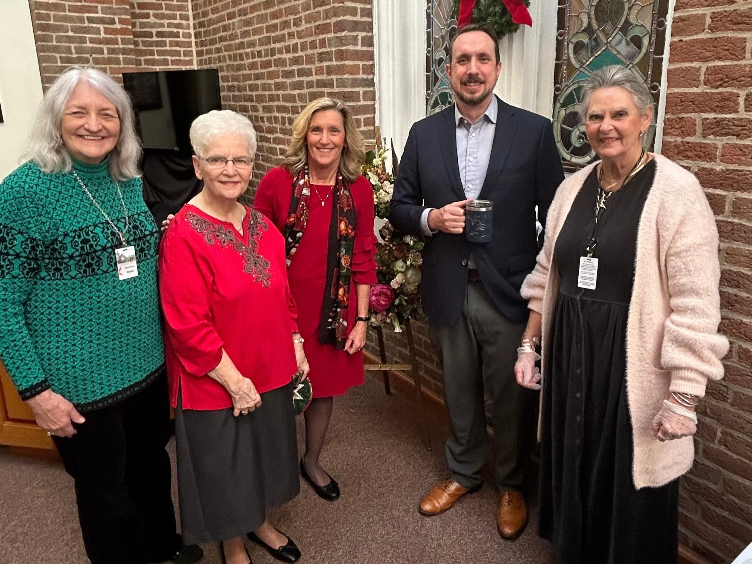 Five people posing in a room with brick walls, some wearing name tags. The group includes four women and one man. The women are dressed in a variety of outfits, including a green sweater and red tops, while the man wears a suit and holds a mug. A decorative stained glass window is visible in the background.