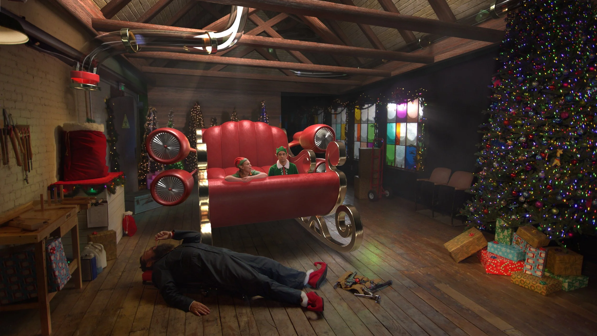 A workshop with a large red sleigh and two people dressed as elves sitting inside. A person lies on the floor with tools scattered around, beside a Santa hat and sack. A decorated Christmas tree with presents is on the right, and holiday lights adorn the area.