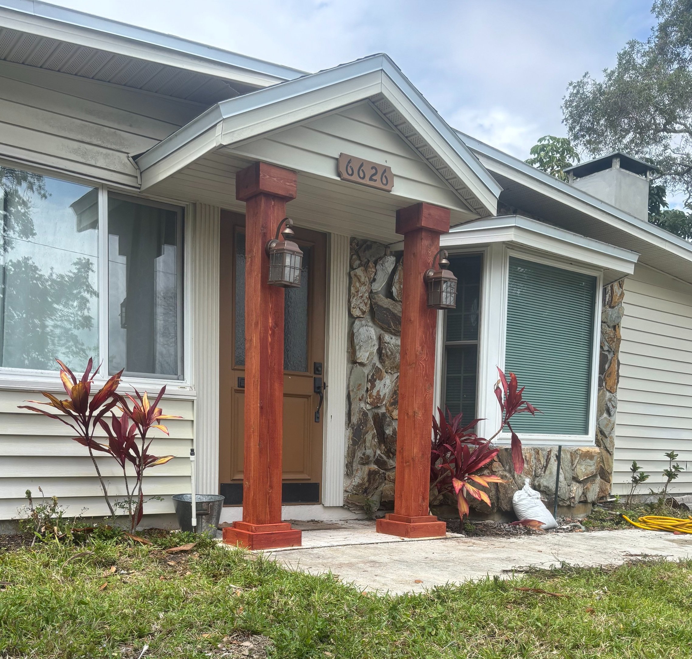 Front entrance of a house with a porch supported by two wooden posts, a brown door, stone and siding exterior, and greenery with plants near the entrance.