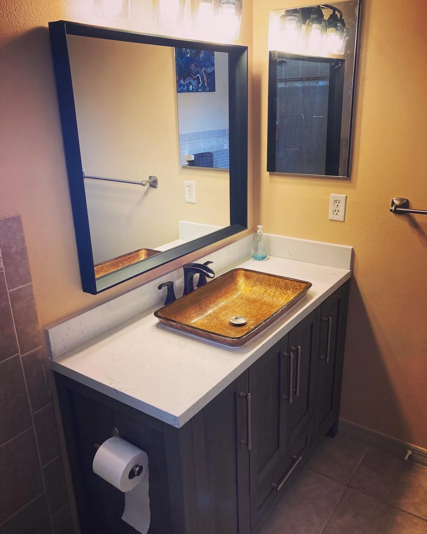 Bathroom vanity with a square golden-brown ceramic sink, a white marble countertop, a black faucet, a mirror, a small bottle of soap, toilet paper, and a wall-mounted cabinet with lights.