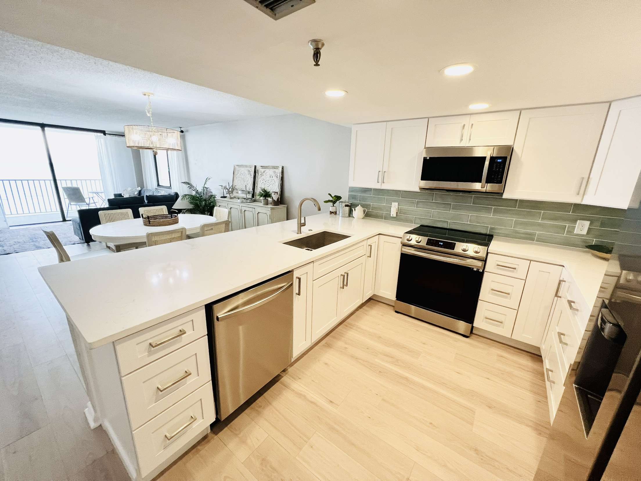 Modern kitchen with white cabinets, stainless steel appliances, light wood flooring, tiled gray backsplash, and an open layout leading to a living and dining area with sliding glass door to balcony.