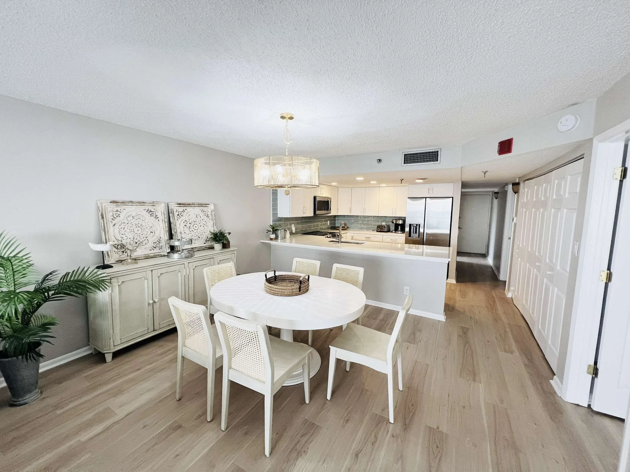 Dining area with white round table, six matching chairs, a sideboard with decorative items, a potted plant, and an open kitchen with white cabinets and stainless steel appliances.
