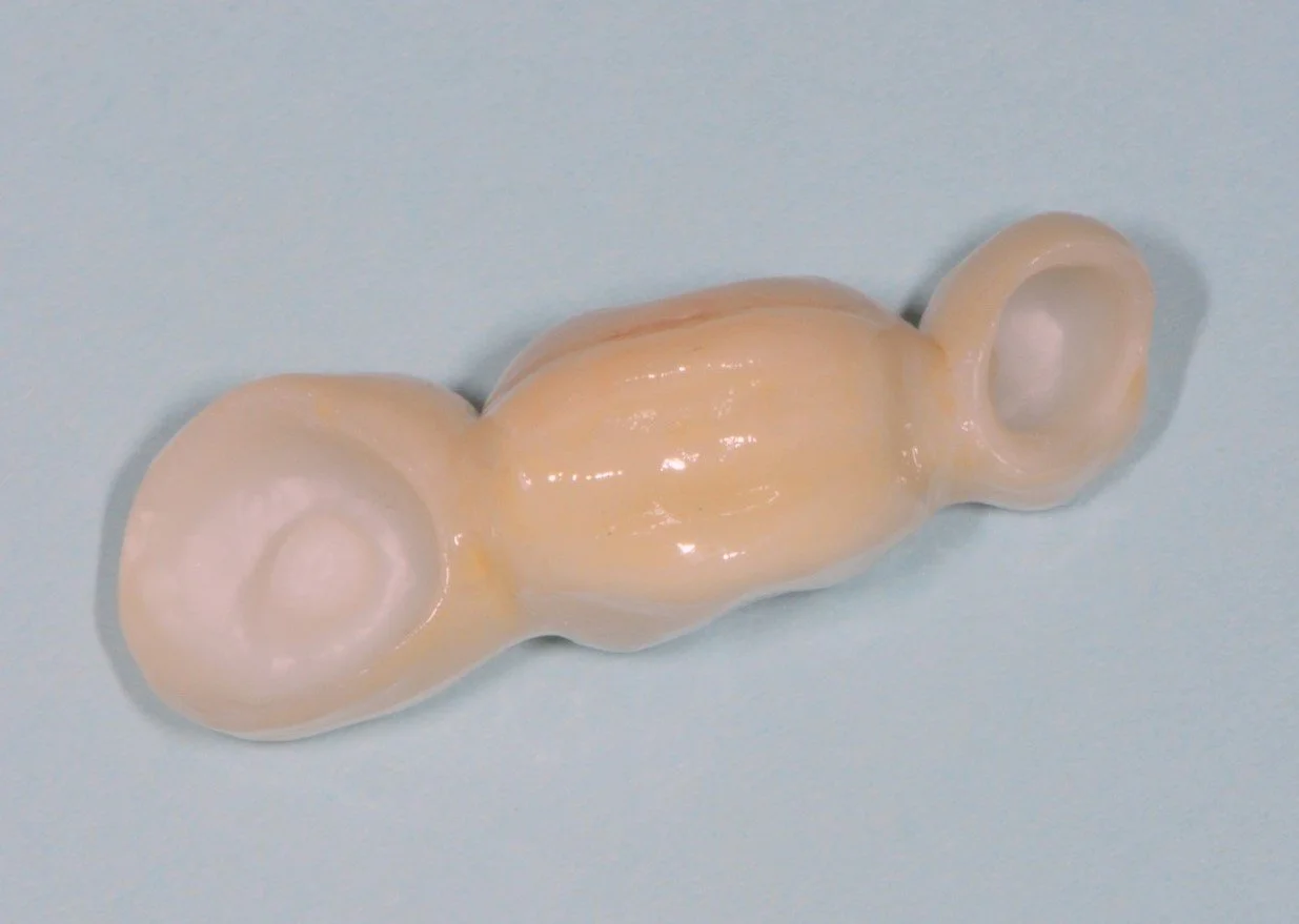 Macro photo of a small, elongated, yellowish ceramic object with two circular ends and a glossy finish, set against a light blue background.