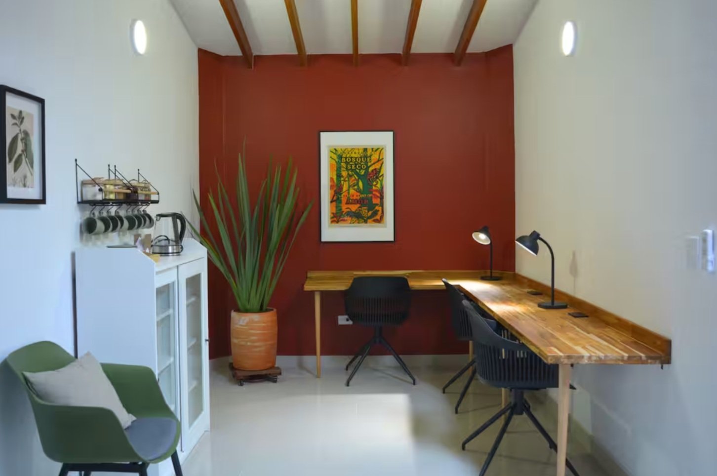 Minimalist office space with two wooden desks, black chairs, and desk lamps. A red accent wall features colorful artwork. A potted plant, green chair, and small shelving unit with mugs and a kettle are on the side.