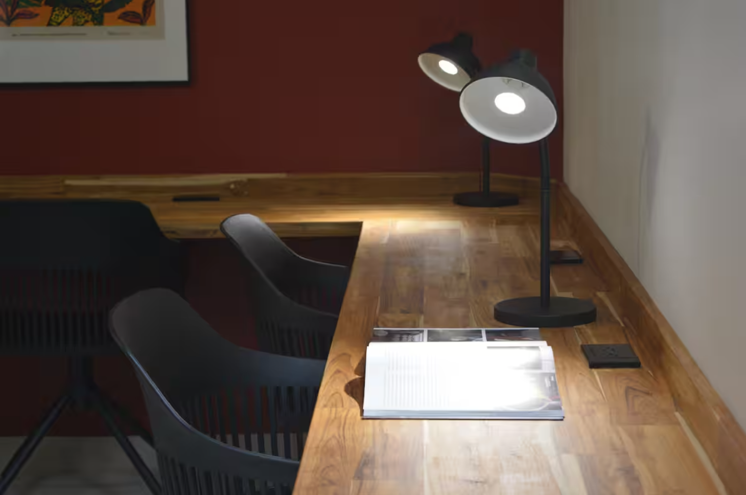 Wooden desk with two black lamps, an open book, black chairs, red wall background, framed artwork
