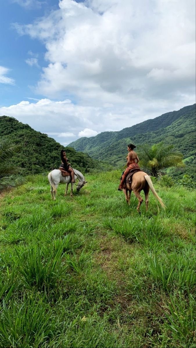 Two people riding horses on a grassy hillside with a scenic view of lush green mountains.