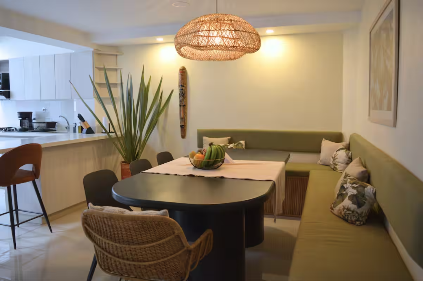 Cozy dining area with green banquette seating, wicker chairs, black dining table, fruit basket centerpiece, woven pendant light, kitchen in background.
