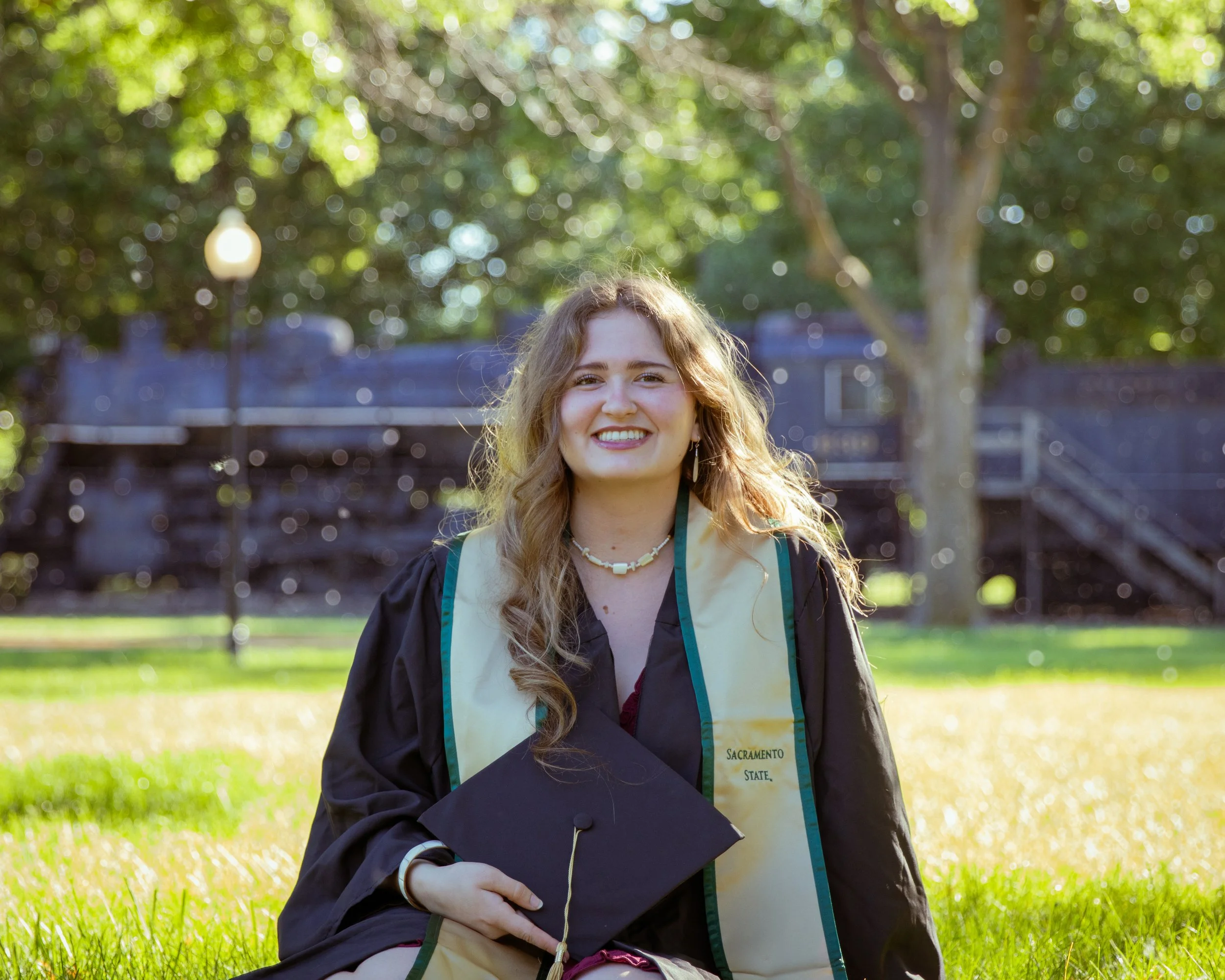 A young woman in a graduation cap and gown sitting outdoors on grass, smiling with a diploma in her hand and a stole that says 'Sacramento State'.