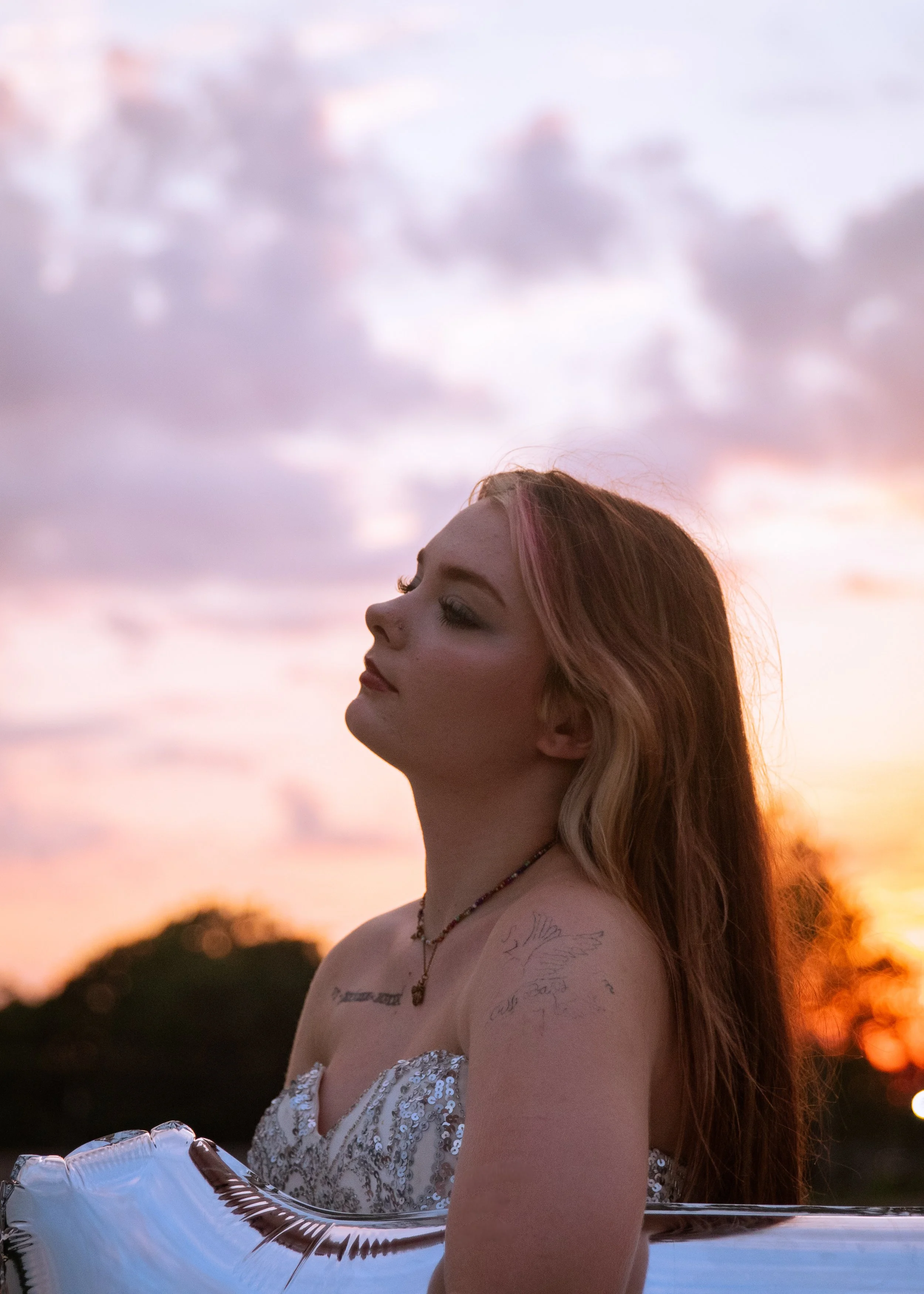 A young woman with long red hair and makeup, wearing a strapless dress, holding a silver balloon, standing outdoors at sunset with a colorful sky and trees in the background.