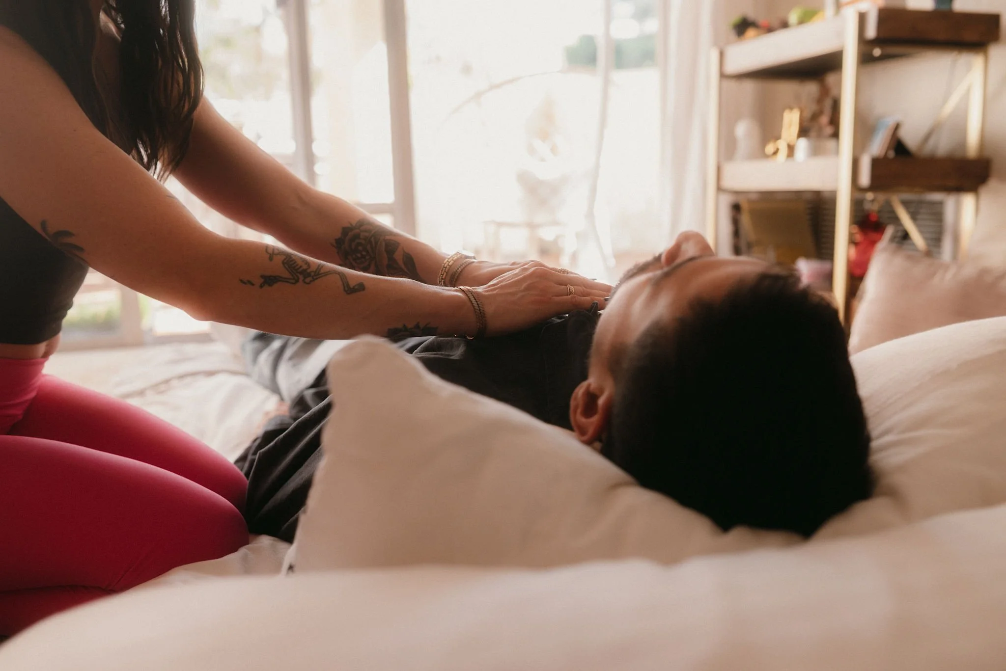 A woman with tattoos on her arms giving a massage to a man lying on a couch in a bright room.