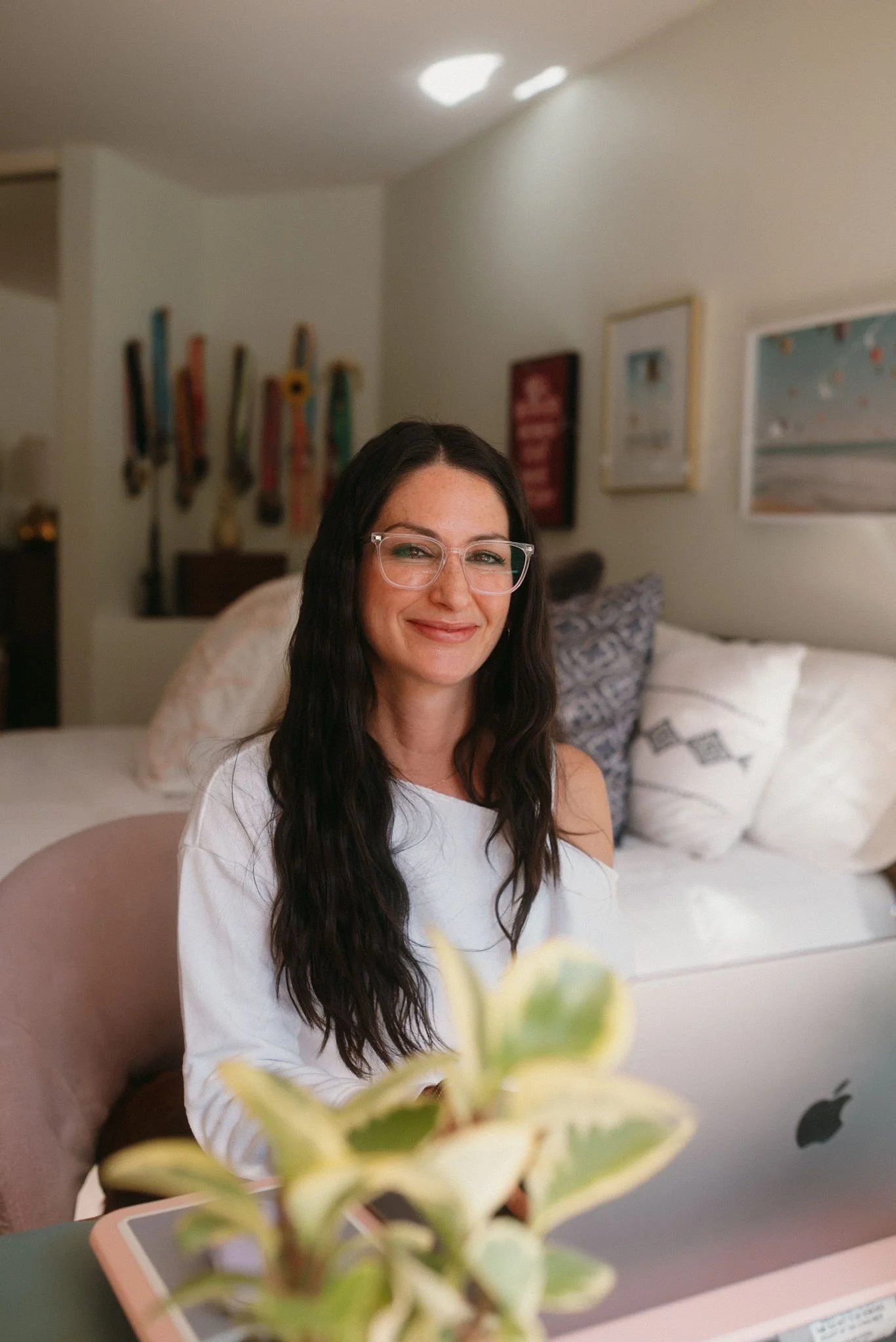 A woman with long dark hair and glasses smiling while sitting on a couch in a living room, with framed pictures on the wall behind her, a plant in the foreground, and a silver laptop on the table in front of her.