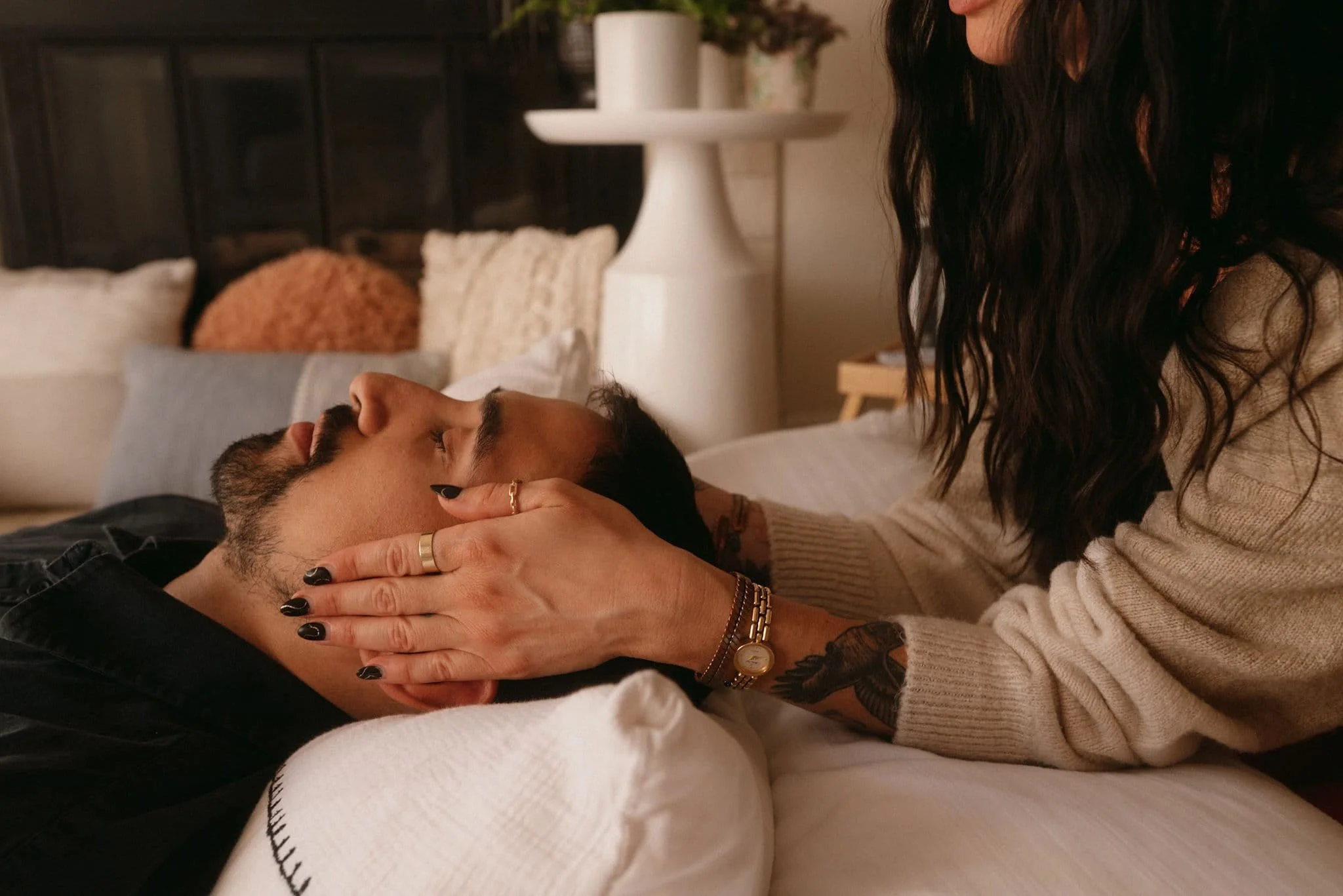 A woman with dark wavy hair and a beige sweater is gently cradling the face of a man with a beard and dark hair, who is lying down with closed eyes. The woman is wearing rings and a watch, and the scene appears to be warm and intimate in a cozy bedroom setting.