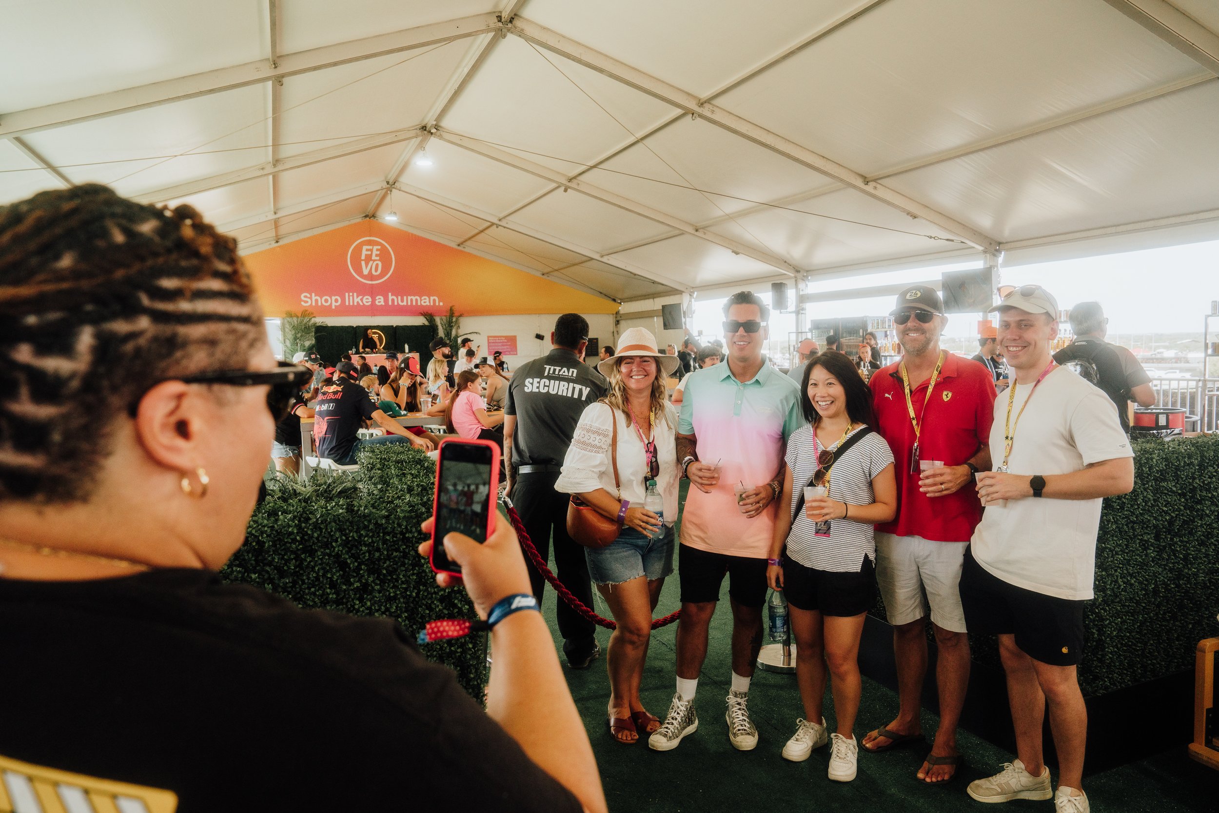 Group of five people posing for a photo at an indoor event with a "Shop like a human" sign in the background. A person is taking the picture on a smartphone. The setting is a tent with other attendees and a security guard visible.