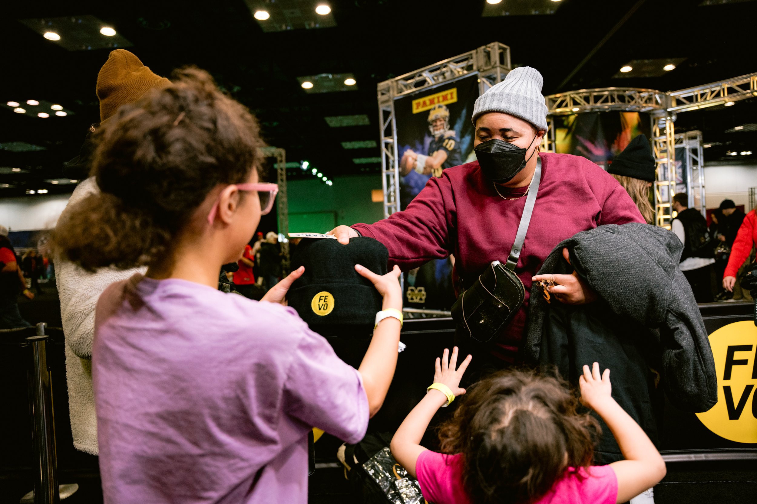 People in a convention center interacting, child receiving a black hat with 'FEVO' logo, purple-shirted person handing it over, background shows crowd and booths.