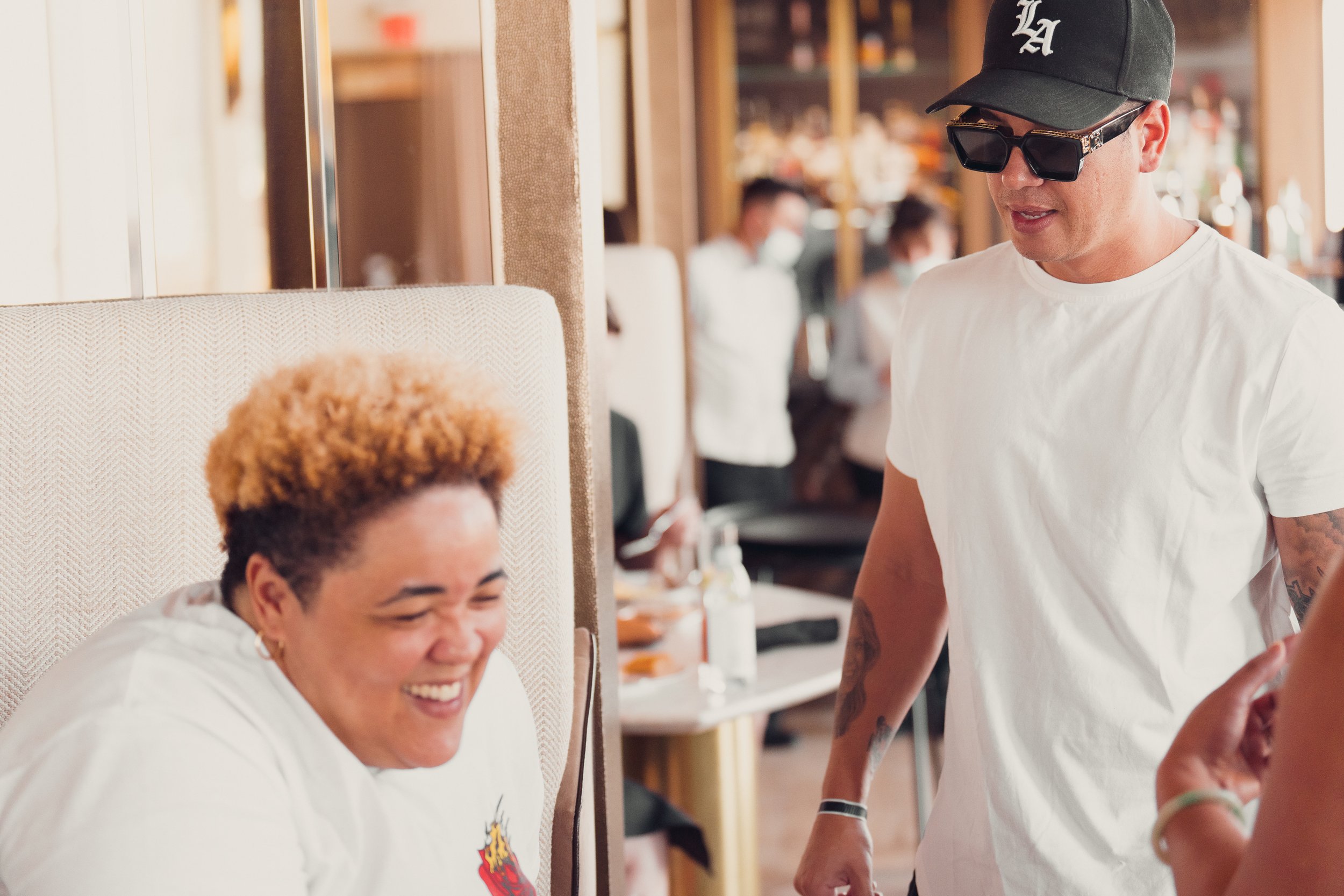 Two people interacting in a lively setting, one smiling broadly while seated, the other standing wearing a white t-shirt and black ball cap, blurred background showing a busy restaurant or café environment.